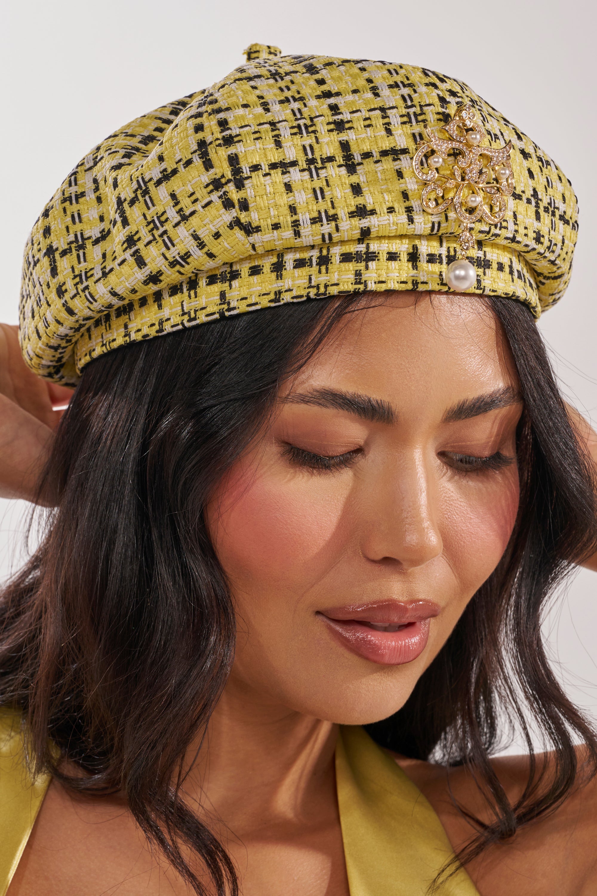 A woman with long dark hair wears the MIA TWEED BERET—Parisian-inspired in yellow and black plaid, adorned with a gold brooch and pearl—while looking down with a gentle expression. 0