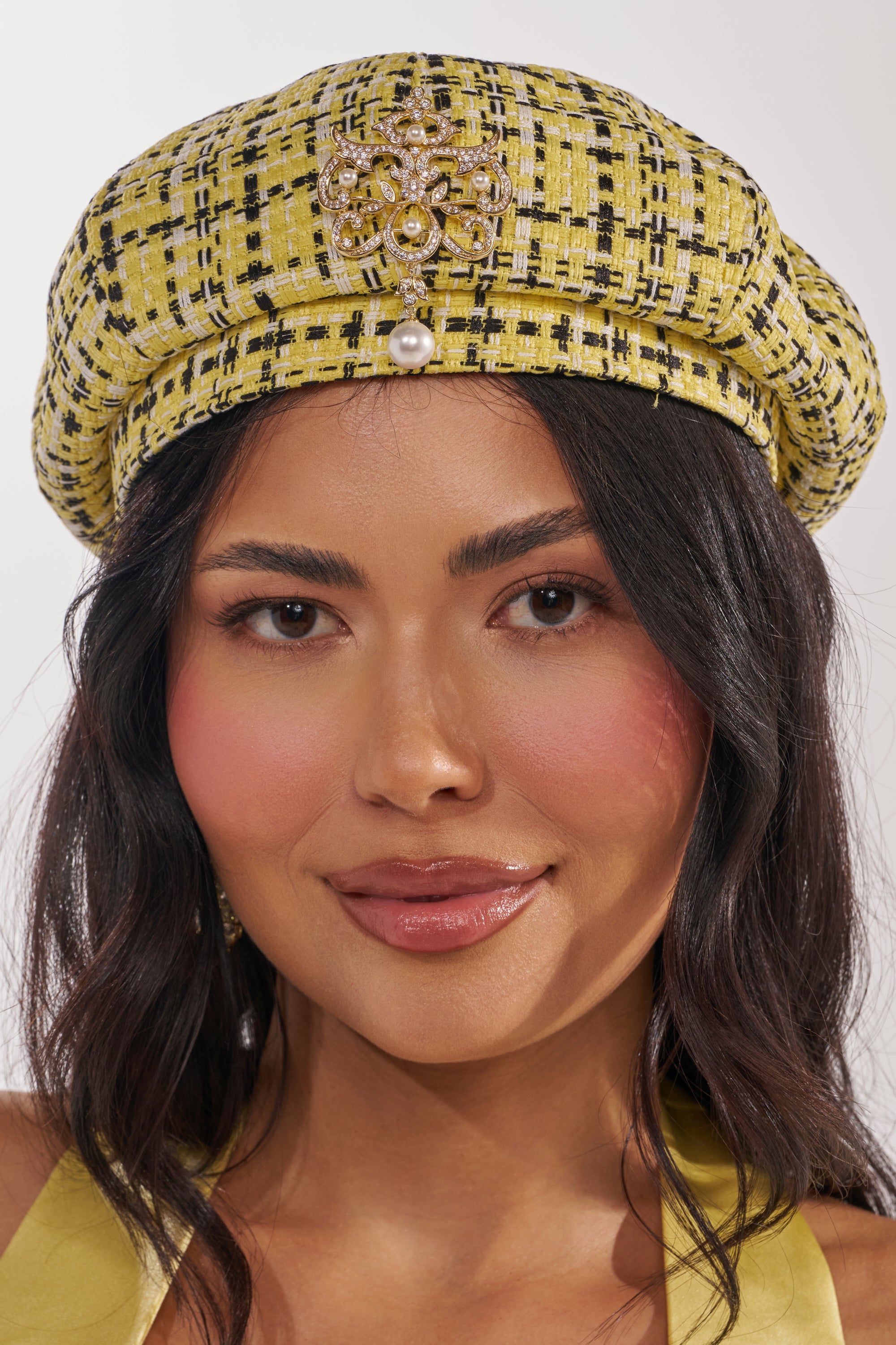 A woman with long, wavy dark hair wears the MIA TWEED BERET, a Parisian-inspired yellow tweed hat featuring a jeweled brooch and pearl drop. She has rosy cheeks, natural makeup, and a slight smile. 2