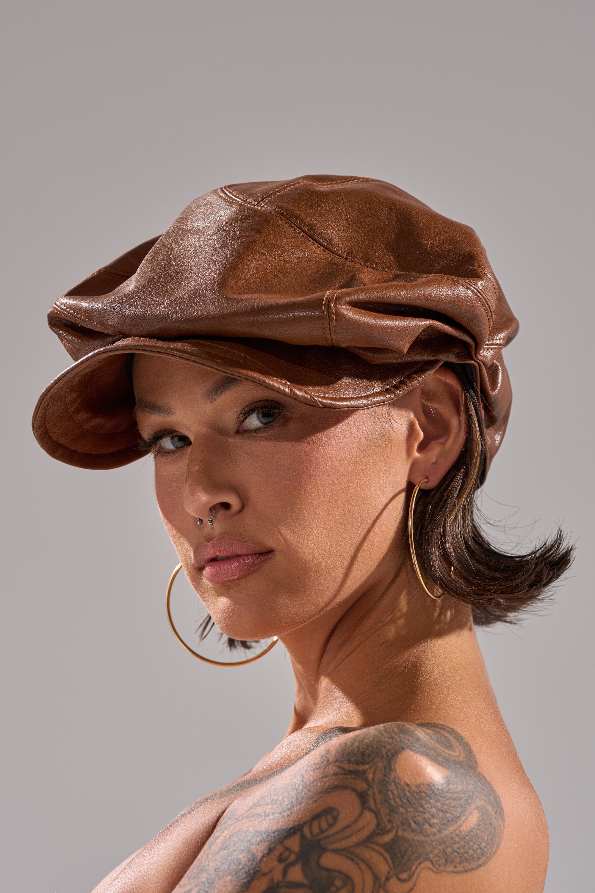 A person with short dark hair wears the PULL IT OFF HAT, gold hoop earrings, and a nose ring. A tattoo is visible on their shoulder as they confidently gaze at the camera, standing against a neutral background.