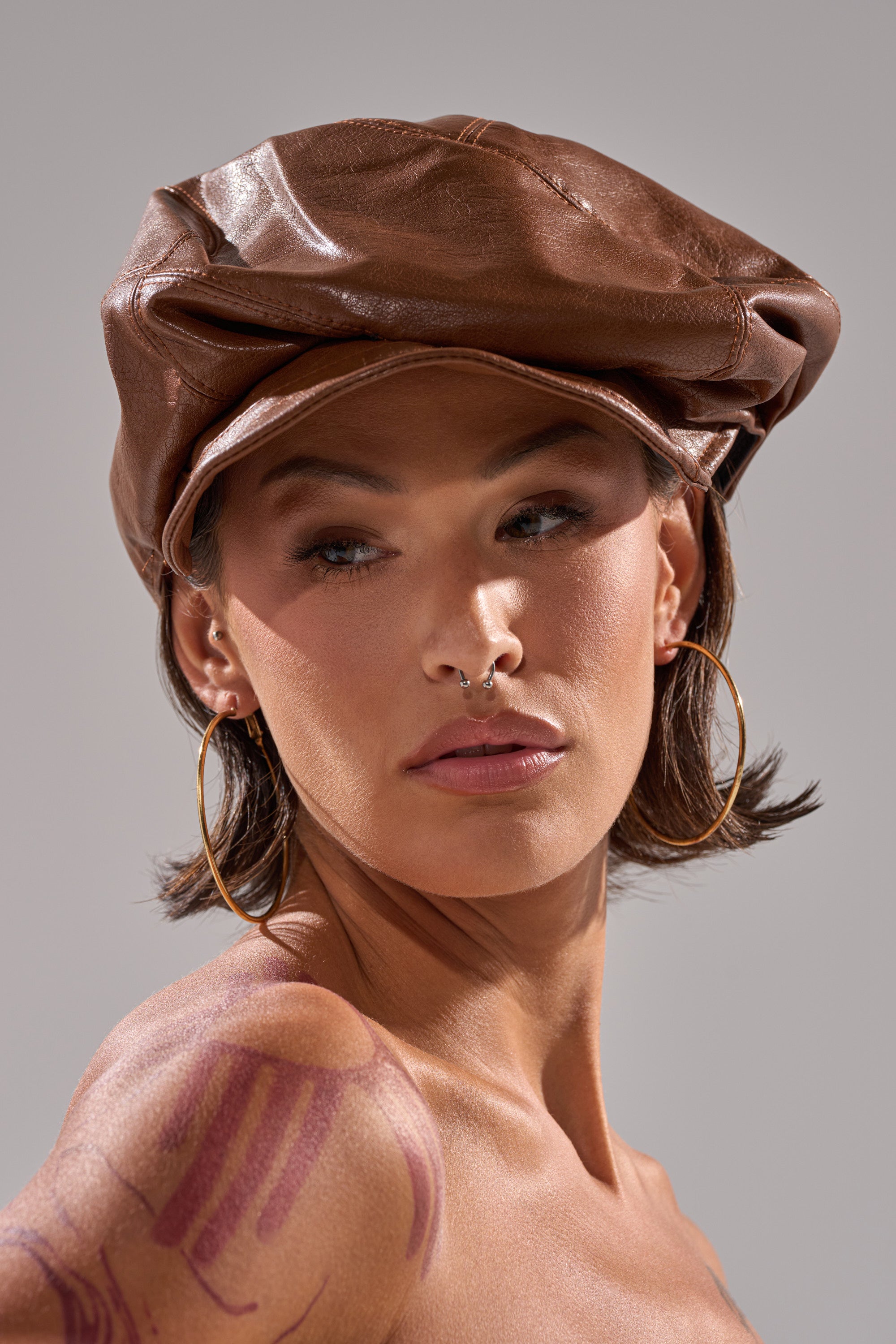 A person with short brown hair wears the PULL IT OFF HAT, large hoop earrings, and a septum piercing, looking to the side. Tattoos are visible on their shoulder as they pose against a neutral background.
