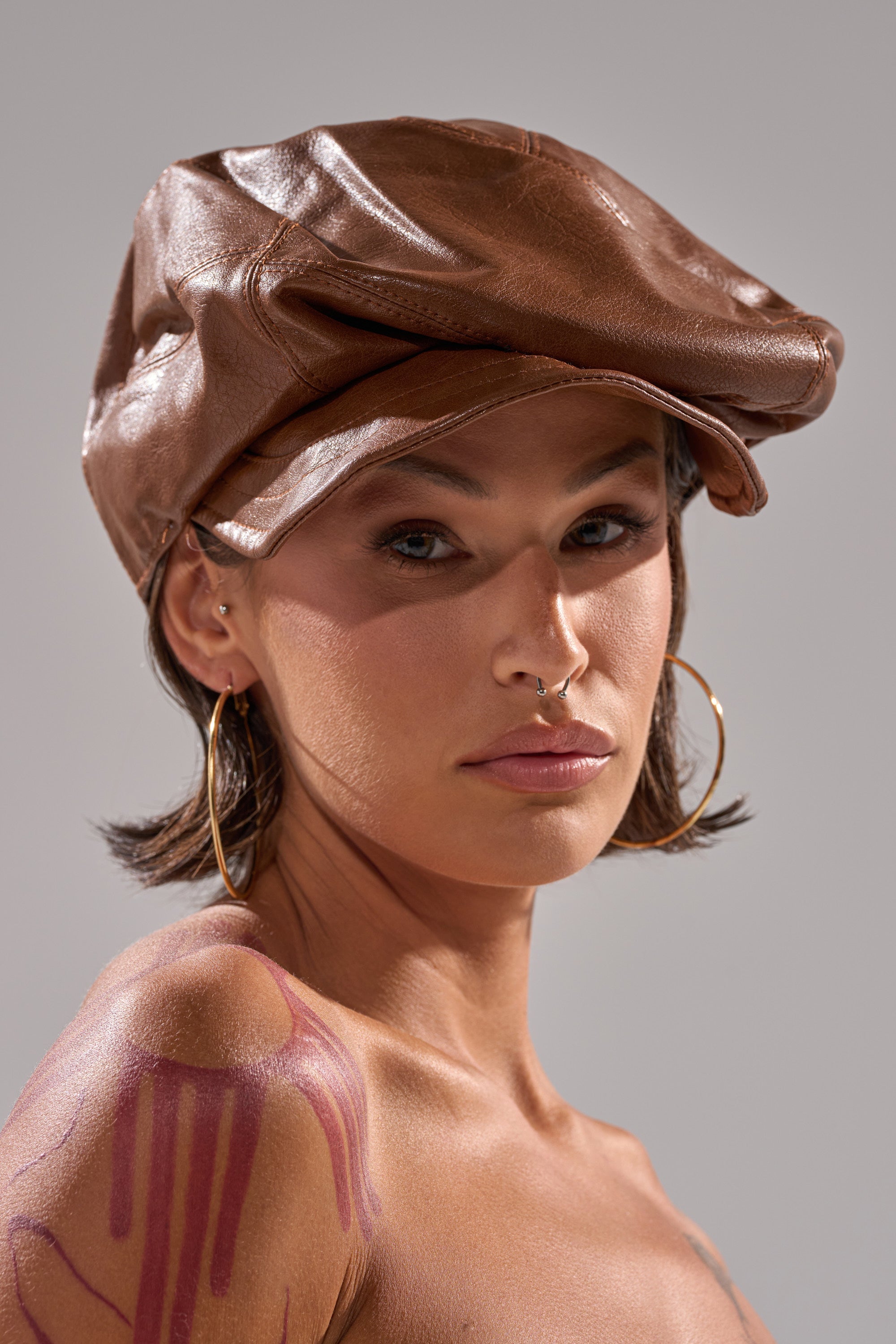 A person with short brown hair wears the PULL IT OFF HAT, large hoop earrings, and a septum piercing while looking at the camera. Artistic painted marks decorate their shoulder against a neutral background. 2