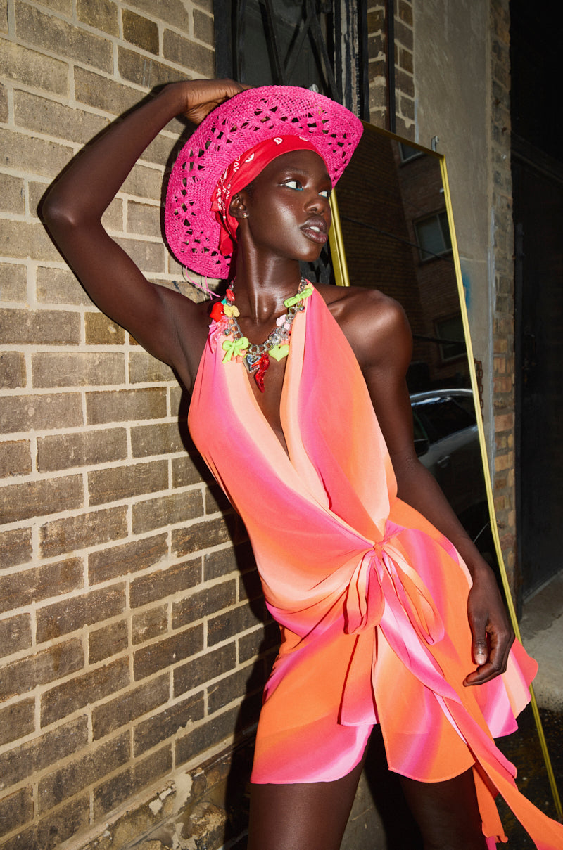 A woman in a vibrant peach and pink halter dress poses confidently against a brick wall, wearing the RIDE EM STRAW COWBOY HAT in bright pink and a colorful statement necklace.