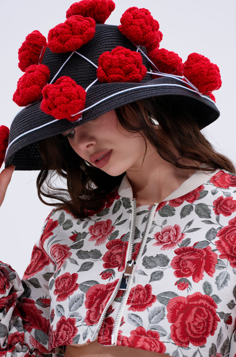 A woman wearing the EPITOME OF LUXURY HAT, a wide brim black hat decorated with red crochet floral accents, and a white and red floral zip-up jacket, looks down while gently touching her hat.