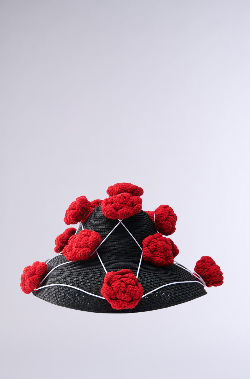 The EPITOME OF LUXURY HAT is a black conical hat adorned with large, textured red pom-poms and white crisscrossing strings, shown on a plain light gray background.