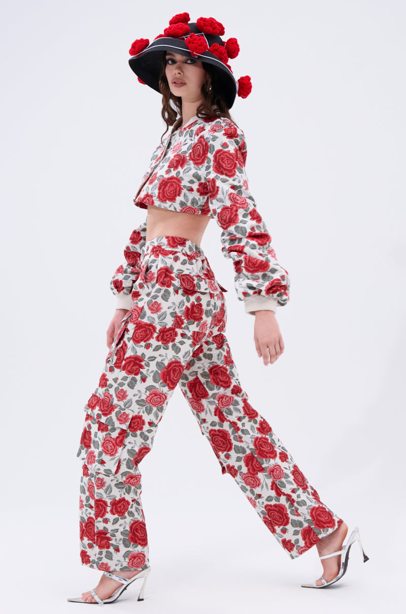 A woman walks in profile wearing a matching red rose print set, white heels, and the EPITOME OF LUXURY HAT—an elegant wide-brimmed hat adorned with large red flowers.