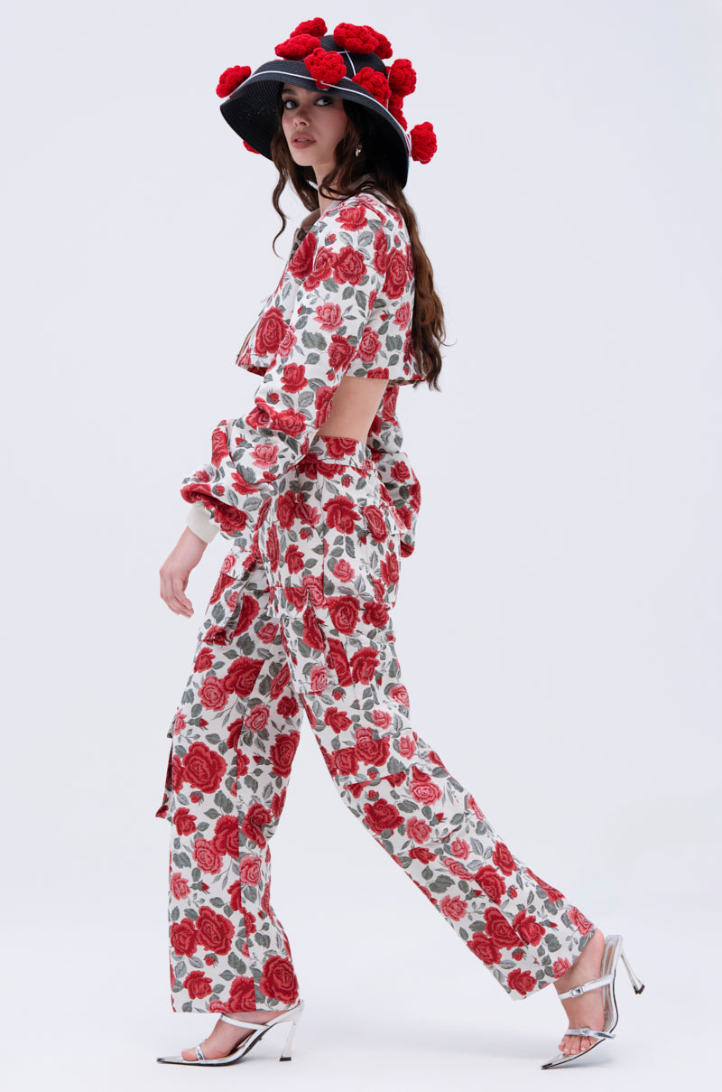 A woman in a matching floral outfit with red roses, featuring a long-sleeve crop top and pants, walks in profile. She wears the EPITOME OF LUXURY HAT adorned with red flowers and silver high heels.