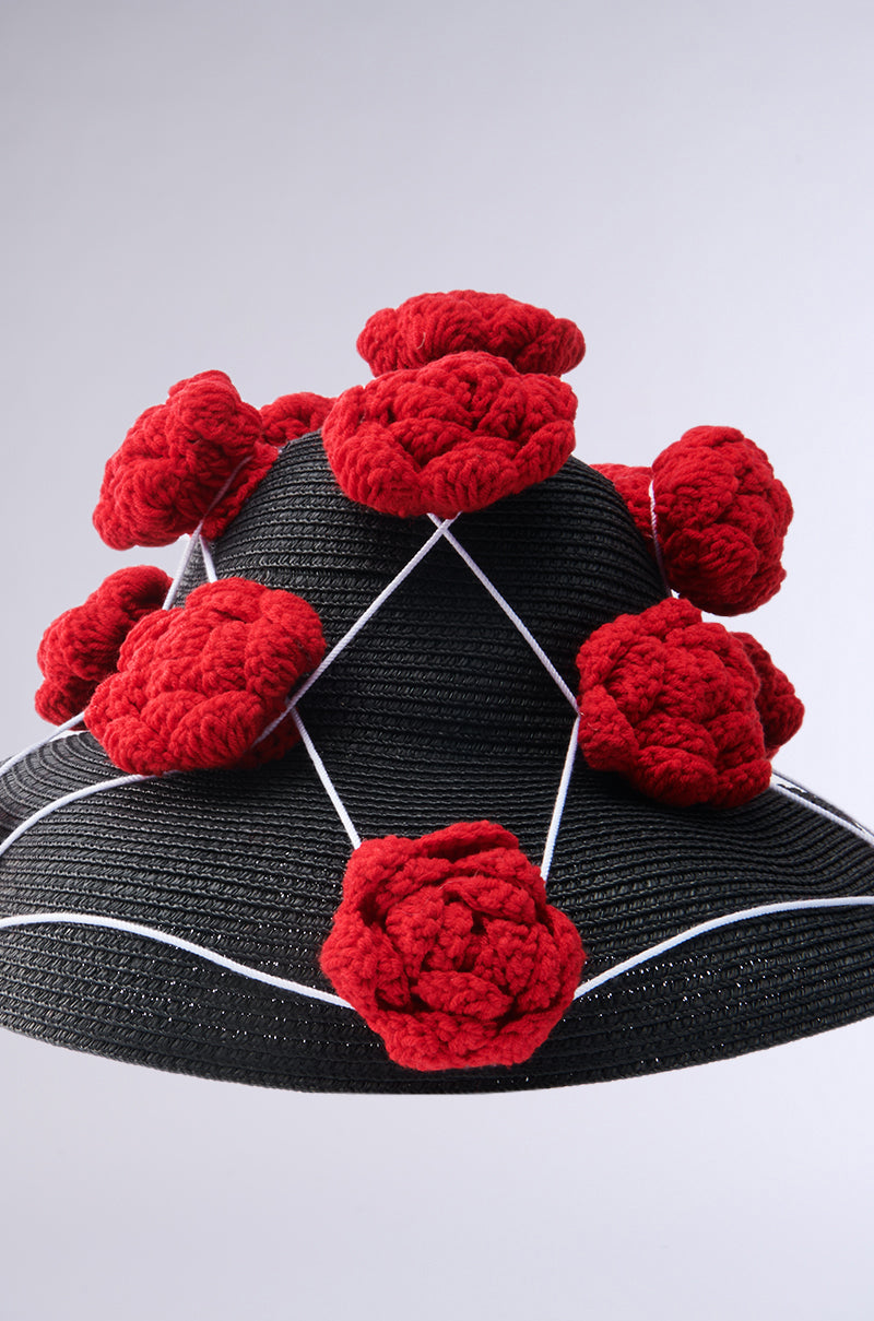 The EPITOME OF LUXURY HAT features a wide brim, red crochet floral appliqués, and white crisscross strings, set against a simple light backdrop.