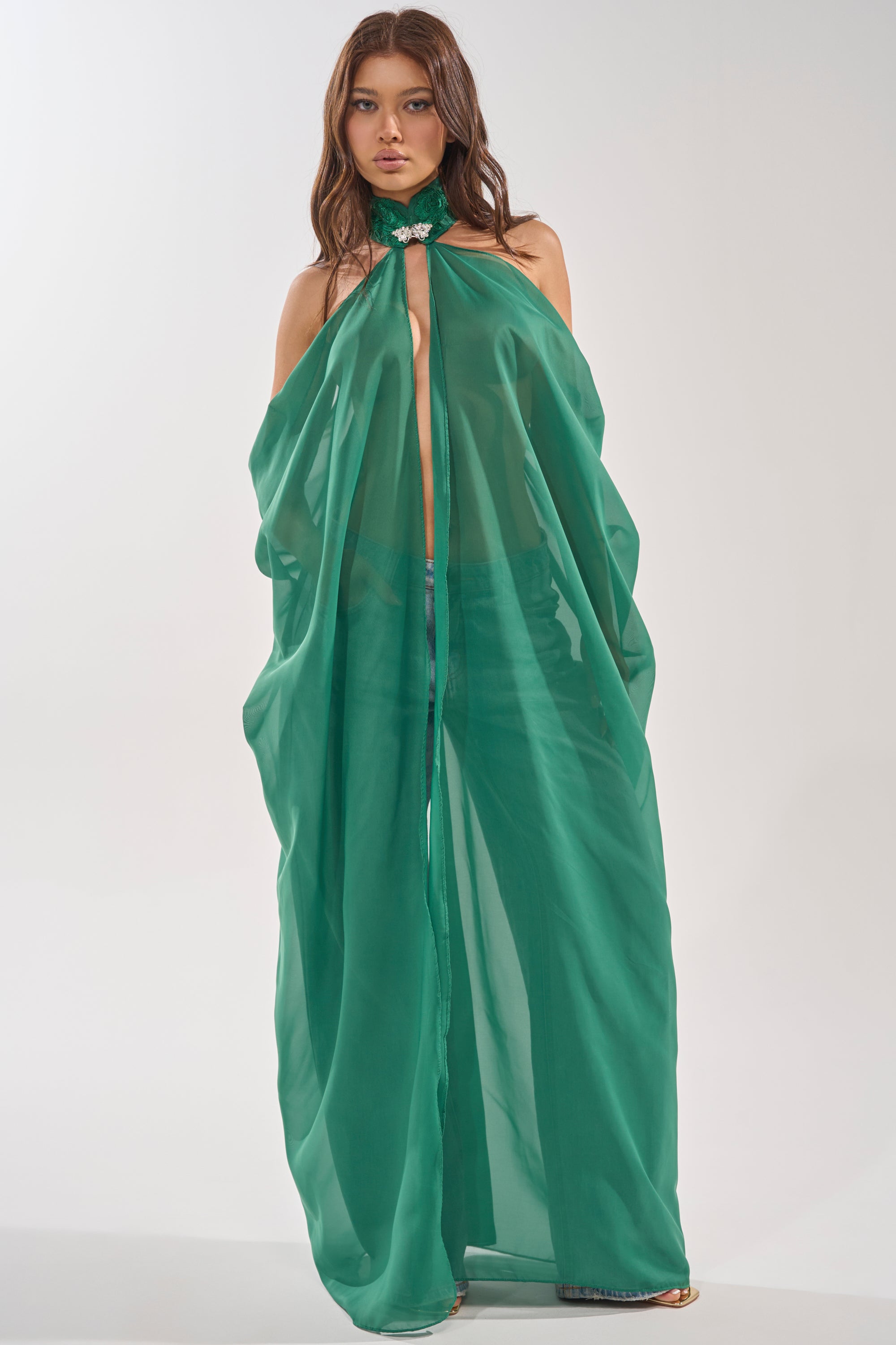 A woman wears the MAKE A STATEMENT CAPE IN GREEN, a sheer green halter-style cape with embellished collar and floral detail. The cape is open at the front, revealing blue jeans. She has long, wavy hair and a neutral expression. 0