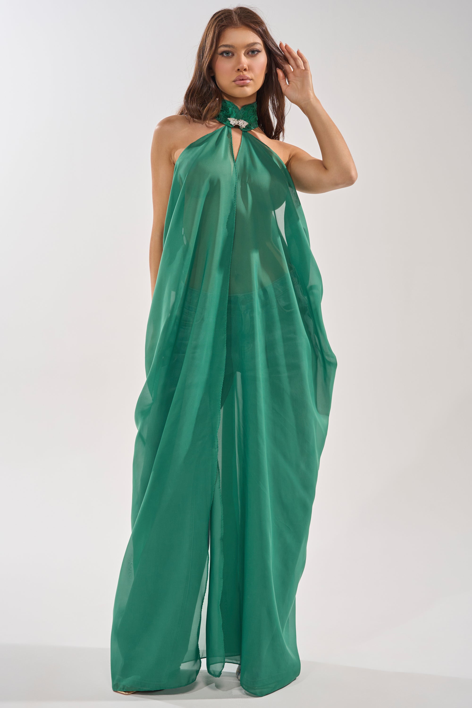 A woman poses confidently in the MAKE A STATEMENT CAPE IN GREEN, featuring sheer, flowing fabric and an embellished collar. The elegant cape drapes gracefully as she stands against a plain background. 1