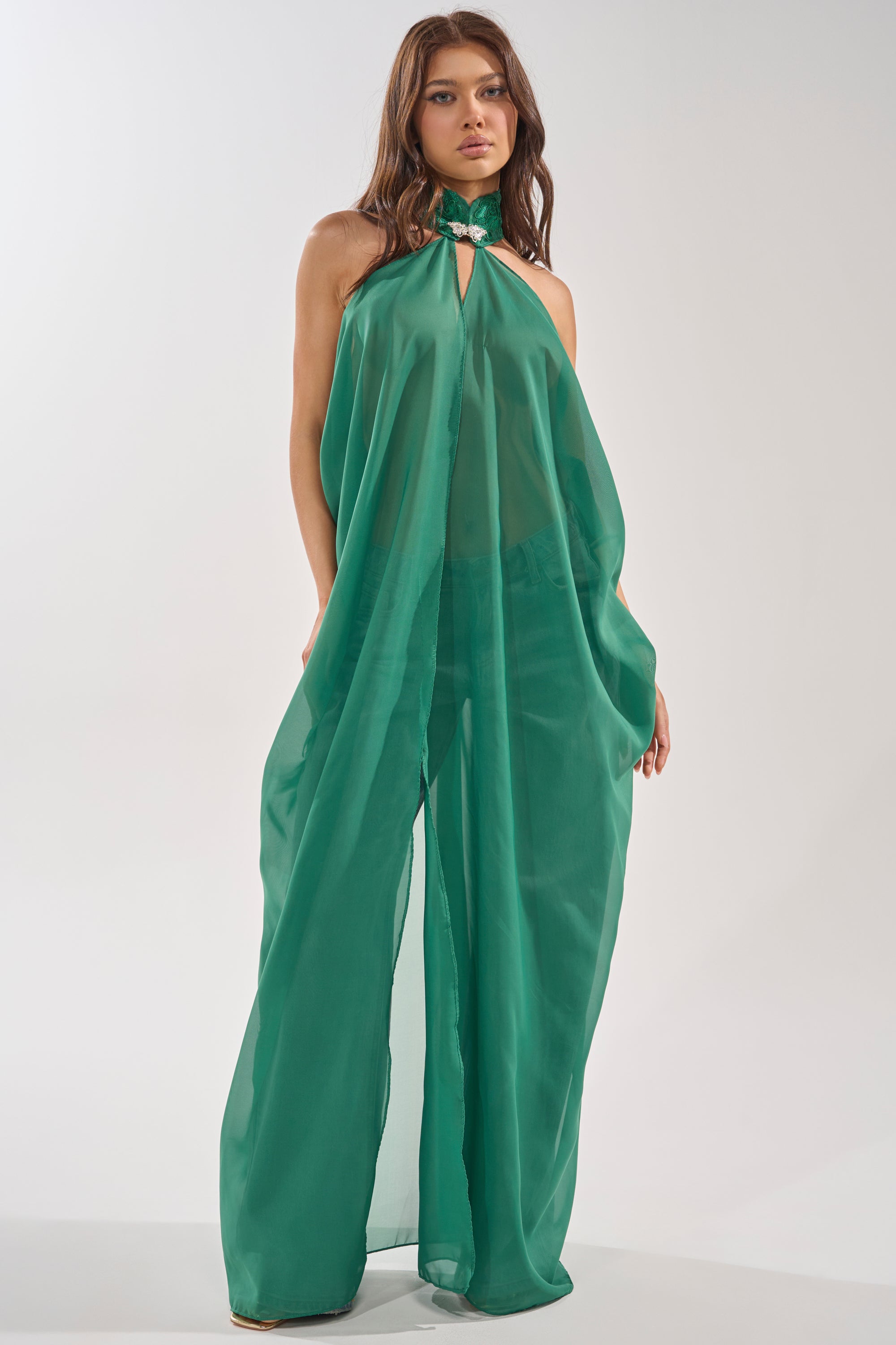 A woman wearing the MAKE A STATEMENT CAPE IN GREEN, a flowing, sheer halter-neck jumpsuit with wide legs and an embellished collar, stands against a plain white background. 2