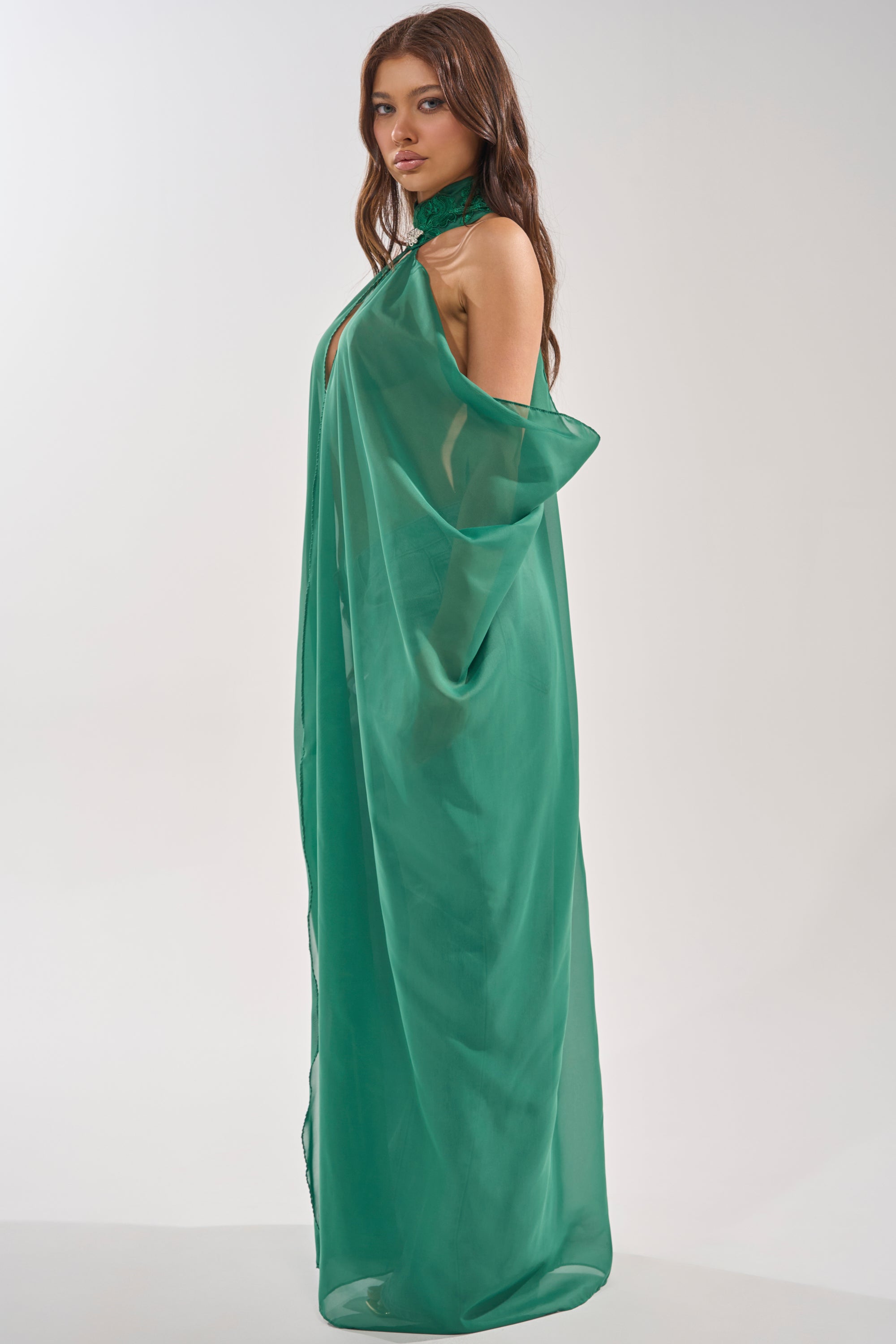 A woman stands in profile wearing the Make a Statement Cape in Green, a loose-fitting, sheer, and elegant piece with an embellished collar. Her long brown hair falls over her shoulders against a plain, light-colored background. 3