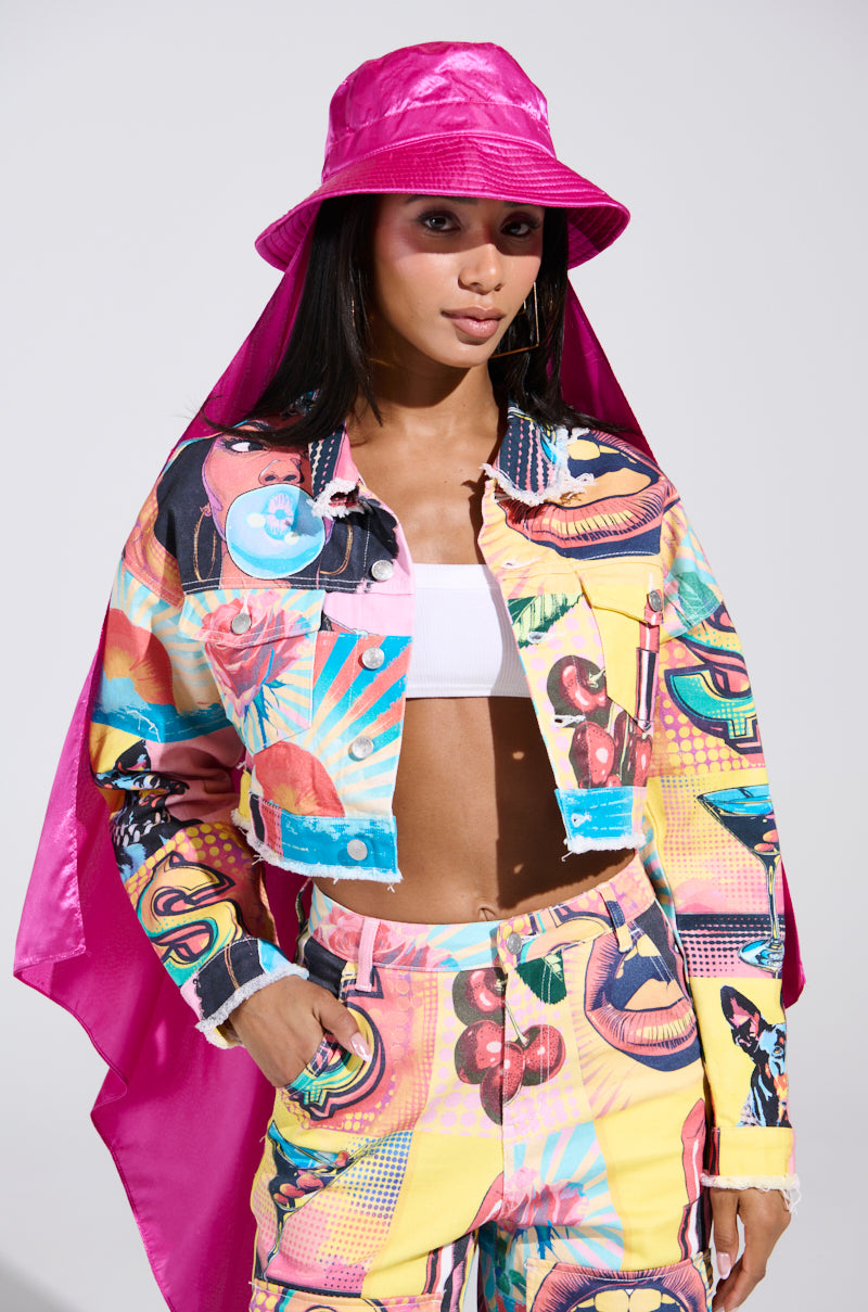 A woman wears a colorful retro-graphic outfit, accented by the HOW YOU DOIN SATIN HAT for sun protection. Her jacket and pants showcase bold, artistic designs over a white crop top, exuding confidence against a neutral backdrop.