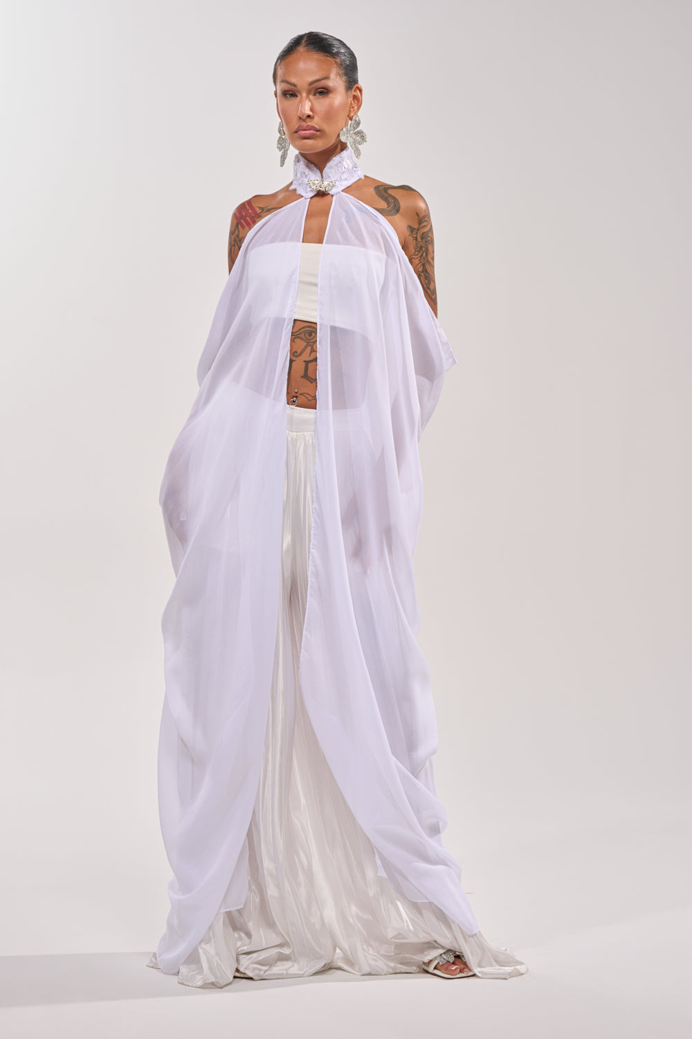 A person stands against a plain backdrop wearing the MAKE A STATEMENT CAPE over a white bandeau top and wide satin trousers, styled with tattoos, statement earrings, an embellished collar, and hair pulled back. 0