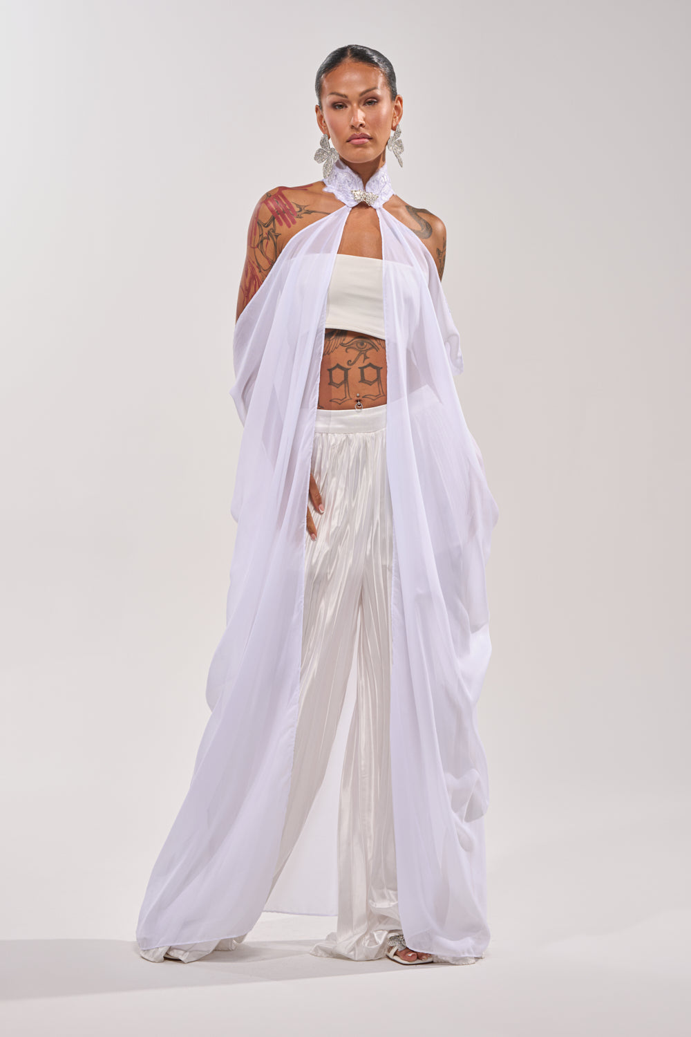 A person stands confidently in the MAKE A STATEMENT CAPE—a flowing sheer organza piece—styled over a white tube top and wide-legged pants, with visible tattoos, statement earrings, and an embellished collar against a plain light background. 1