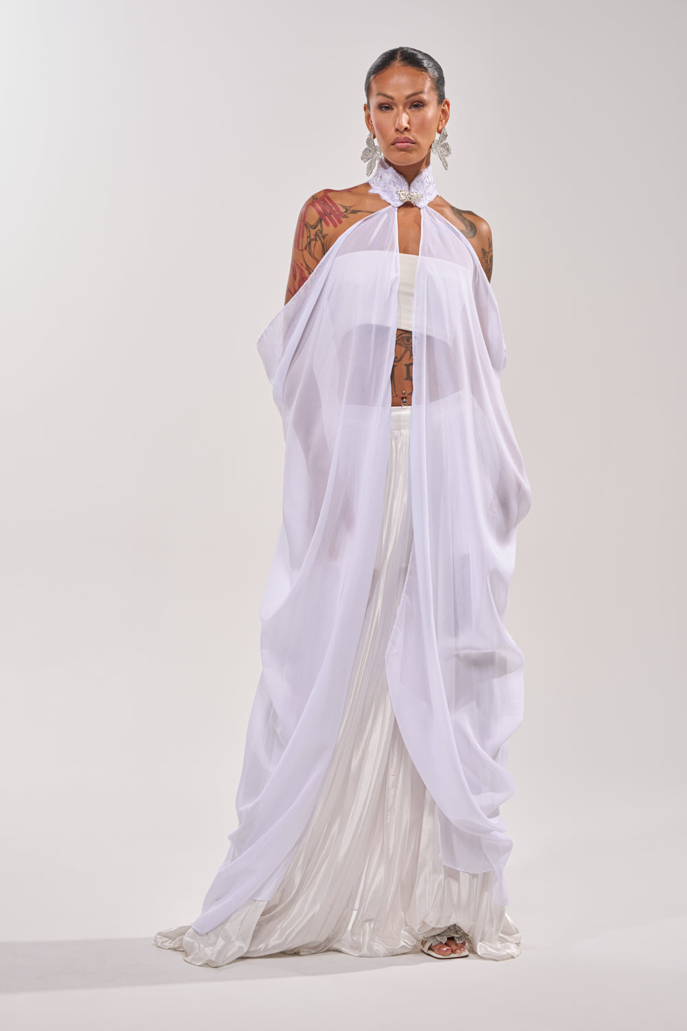 Person stands against a plain background, showcasing tattoos while wearing the MAKE A STATEMENT CAPE—a sheer organza piece over a flowing white skirt—styled with large earrings and an embellished collar for a chic formal look. 2