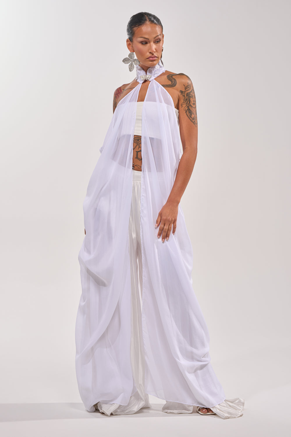 A woman models the MAKE A STATEMENT CAPE—a sheer white halter-neck outfit with wide-legged pants—paired with bold silver earrings and a choker, posing against a plain light backdrop. 3