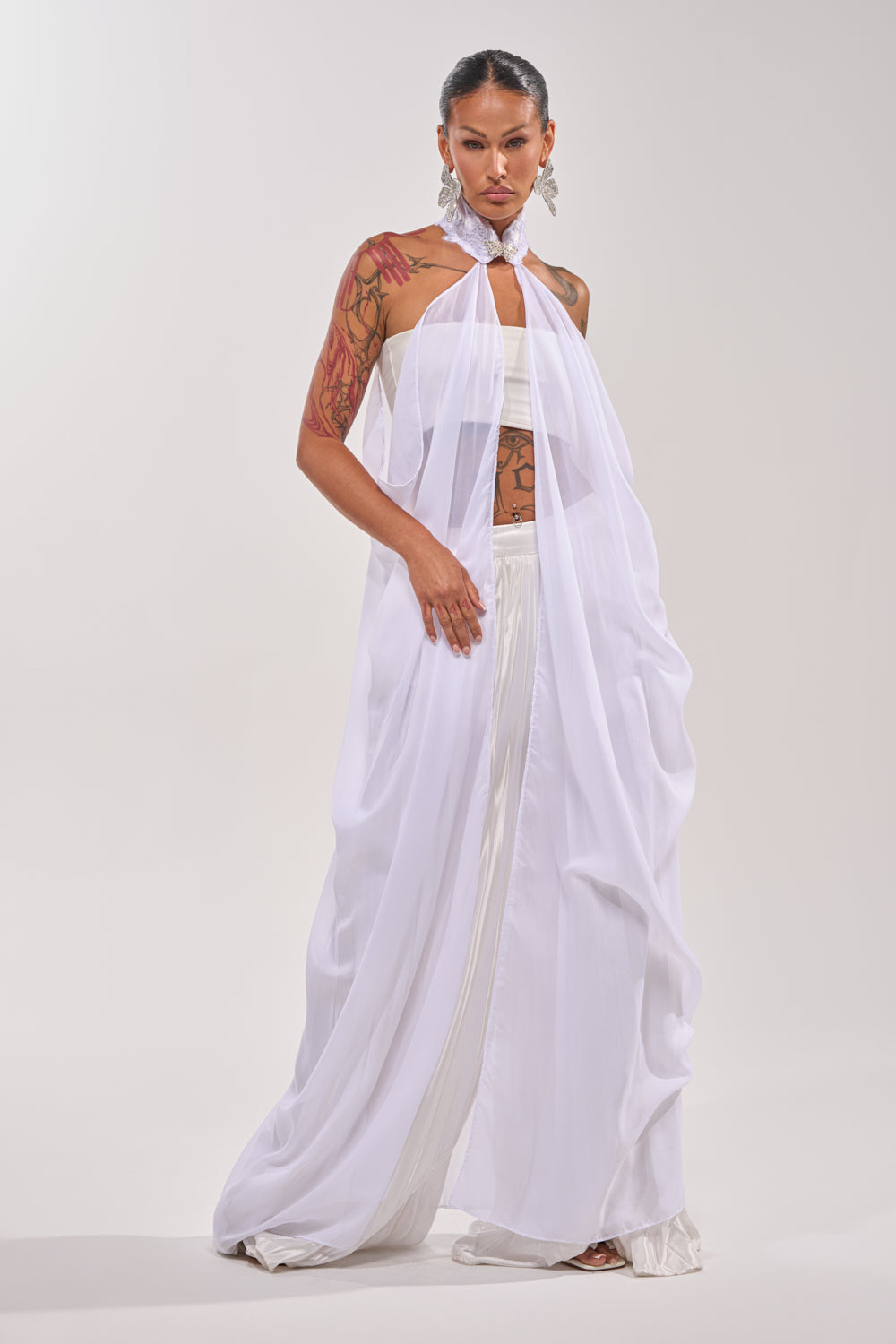 Wearing the MAKE A STATEMENT CAPE, white pants, and a strapless top, a person with visible arm tattoos, bold earrings, and sleek hair exudes elegance against a simple light background. 4
