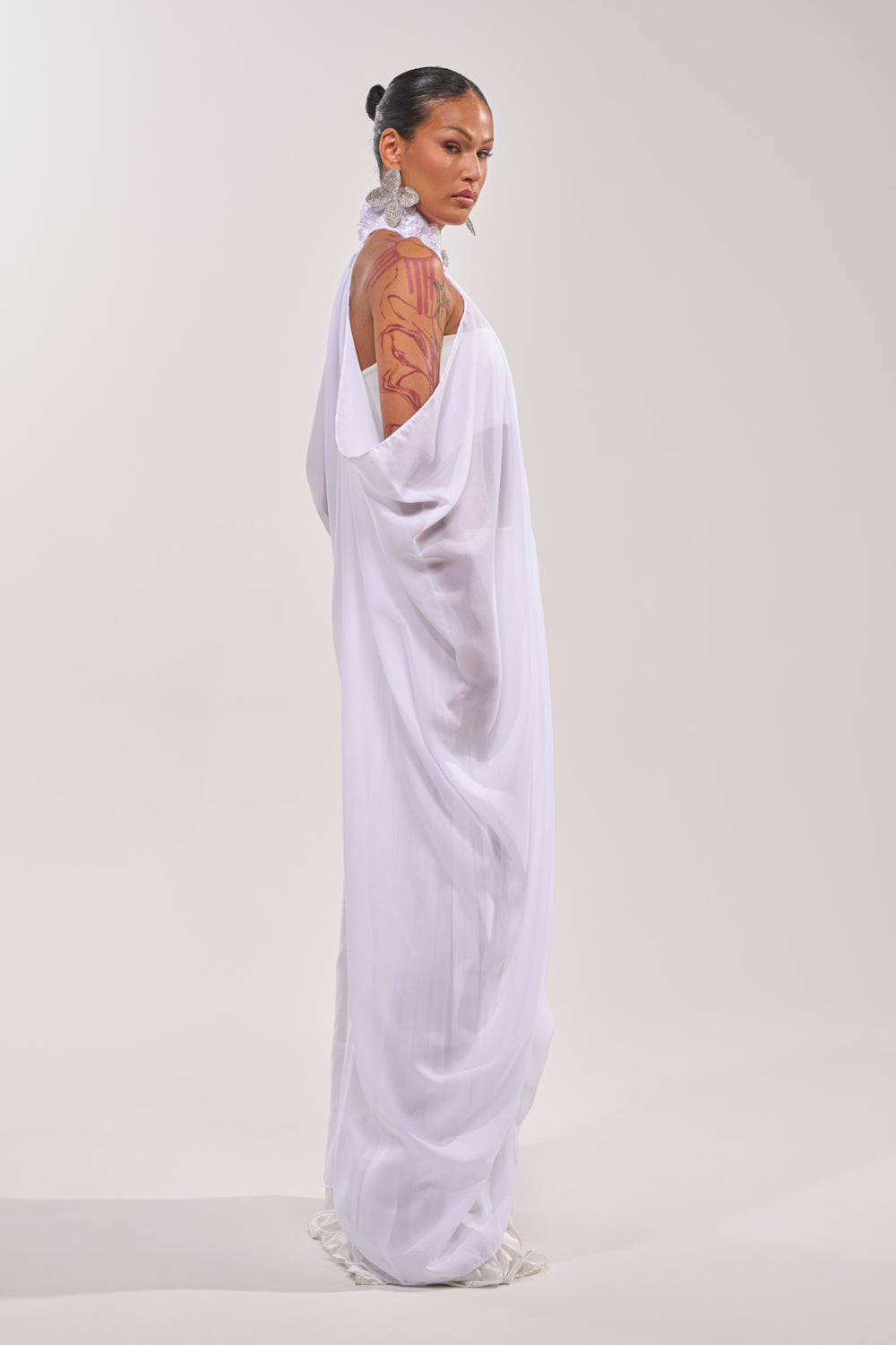 A woman in profile wears a flowing sheer white dress paired with the MAKE A STATEMENT CAPE. Her hair is slicked back, she sports large floral earrings and bold red arm art, against a simple light backdrop. 5