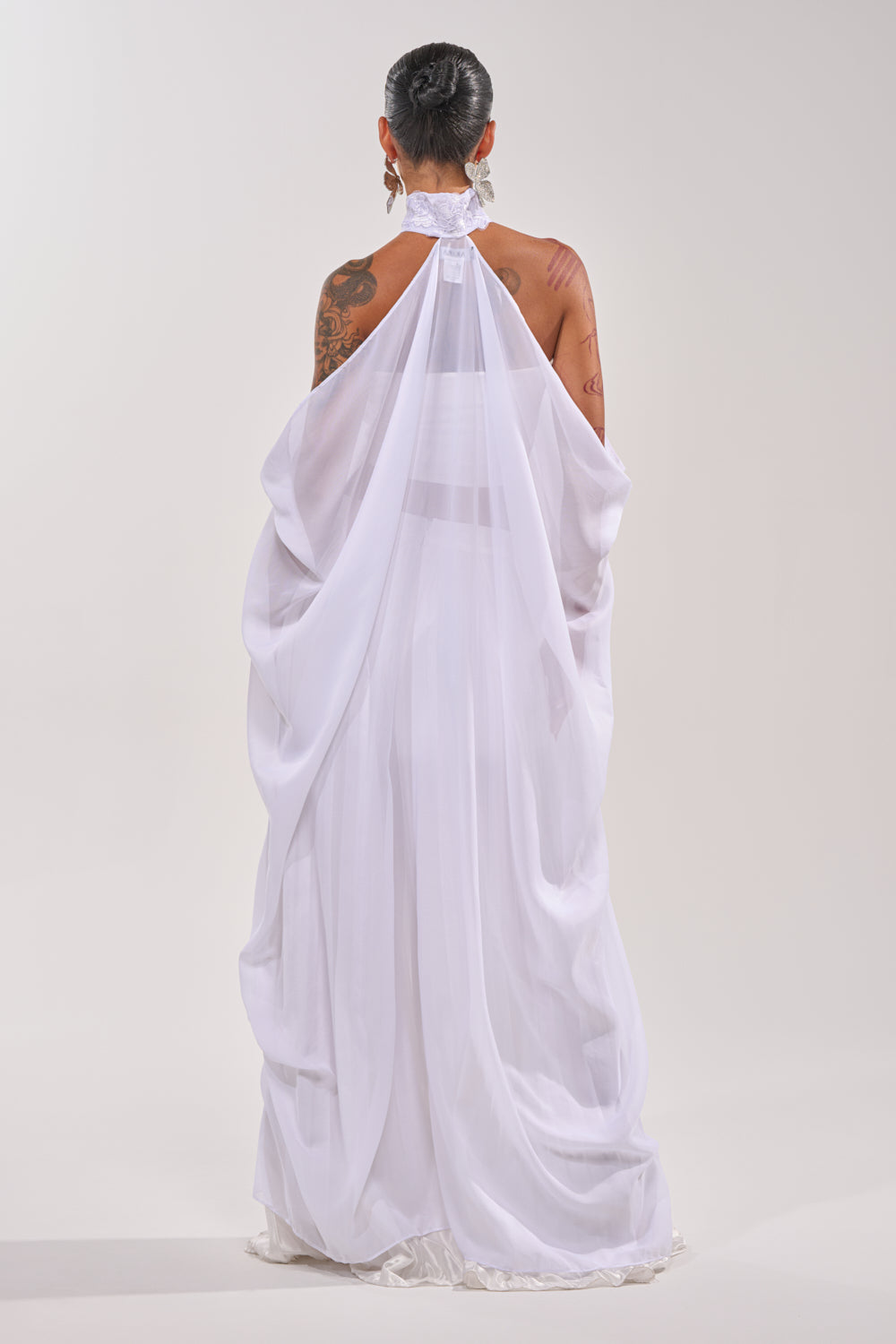 A person with visible tattoos, hair in a neat bun, wears a flowing sheer white halter-neck dress paired with the MAKE A STATEMENT CAPE. They stand facing away against a plain light background, highlighting the cape’s elegant, formal style. 6