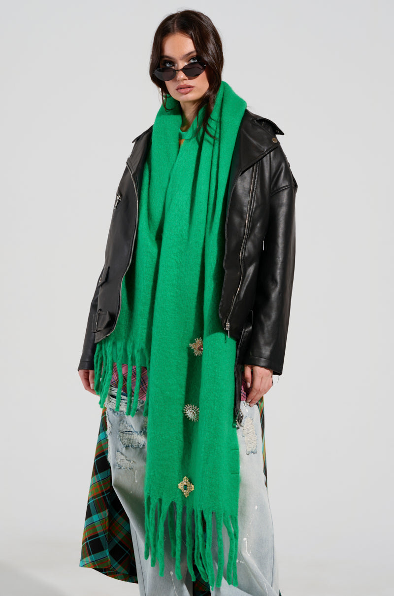 A person wearing the "Embellish Me" brooch scarf in a long green style adorned with gemstones, paired with a black leather jacket, oversized jeans, and sunglasses poses against a plain background. The ensemble is accentuated by a plaid hem. 1