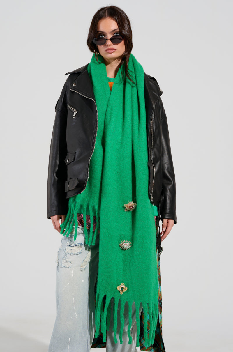A person wearing an EMBELLISH ME BROOCH SCARF in green, featuring gemstone embellishments, paired with a black leather jacket, distressed light blue jeans, and sunglasses. The scarf is long with fringed ends, set against a plain white background. 2