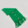An EMBELLISH ME BROOCH SCARF with fringed edges rests on a white surface, featuring three colorful embroidered floral designs, making it a stylish winter accessory.
