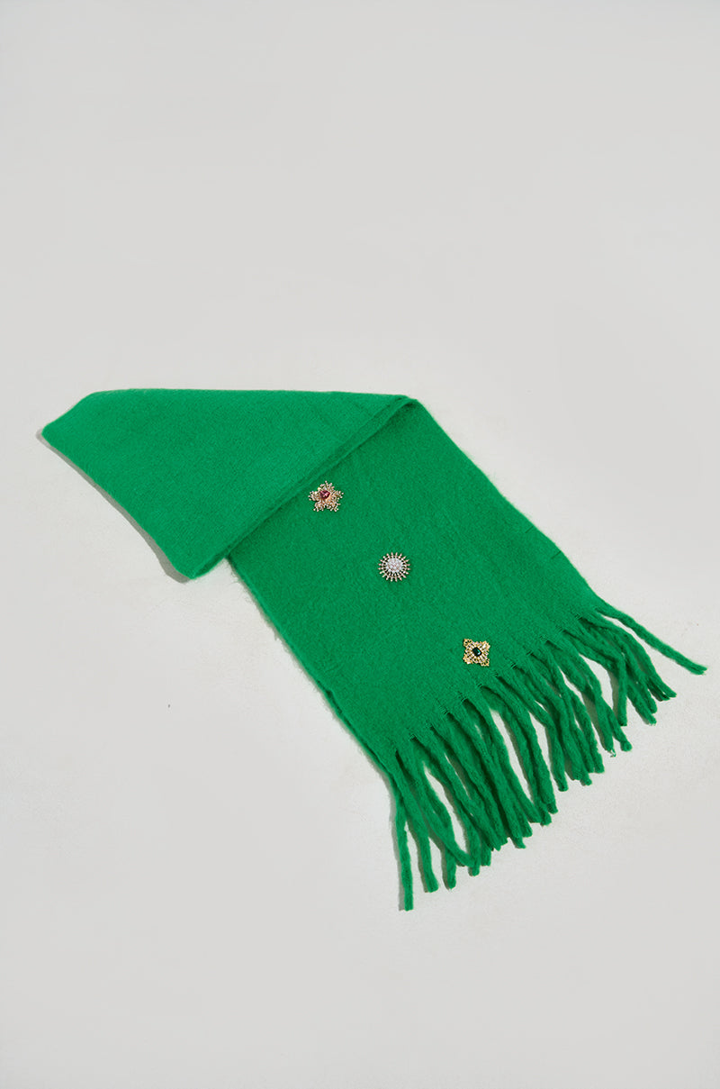 An EMBELLISH ME BROOCH SCARF with fringed edges rests on a white surface, featuring three colorful embroidered floral designs, making it a stylish winter accessory. 0