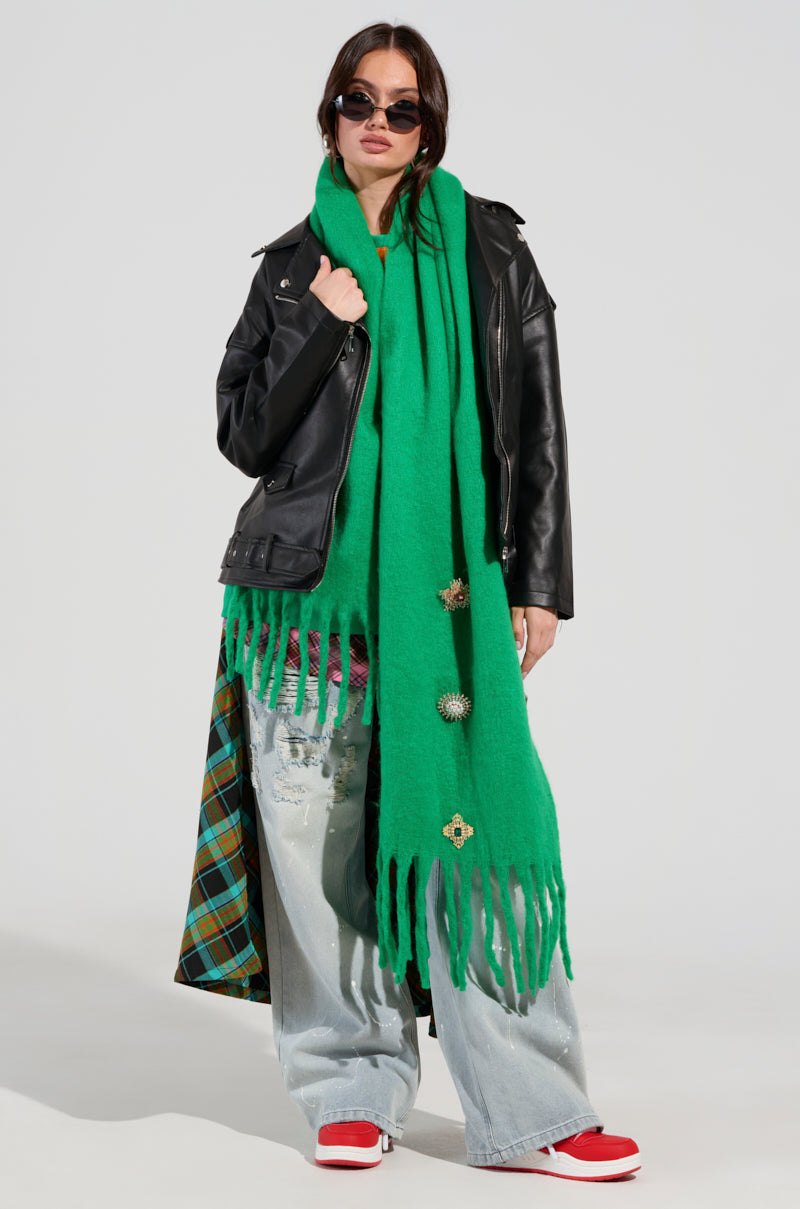 A person wearing sunglasses and the Embellish Me Brooch Scarf with fringe, stylishly adorned with gemstone embellishments, pairs it with a black leather jacket, denim jeans, and red sneakers, standing confidently against a plain background. 4