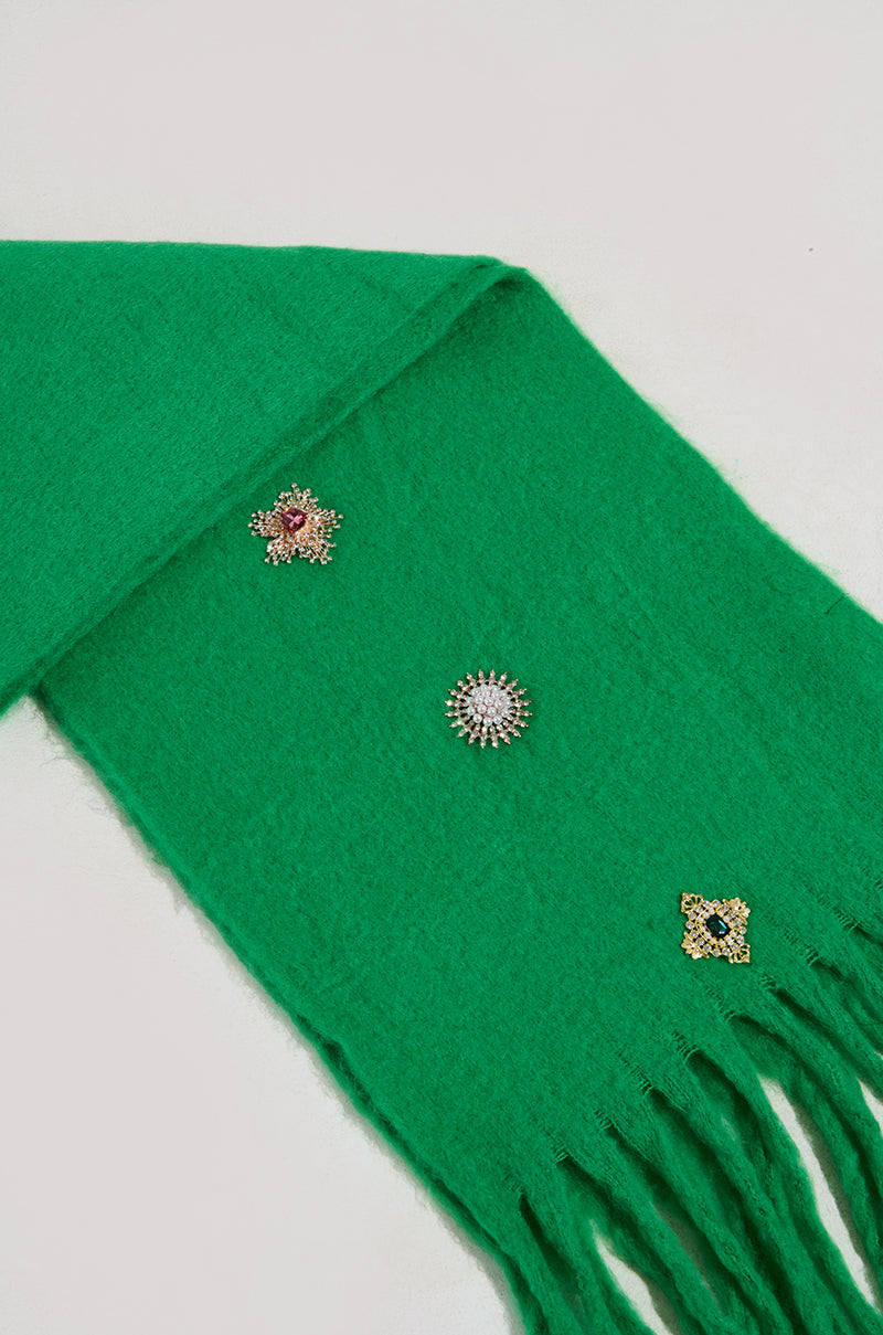 The EMBELLISH ME BROOCH SCARF is a stylish winter accessory in green, highlighted with fringed ends and adorned with three decorative brooches: a pink flower, a round design set with clear stones, and a yellow flower featuring a pink center. It is elegantly showcased against a light gray background. 3