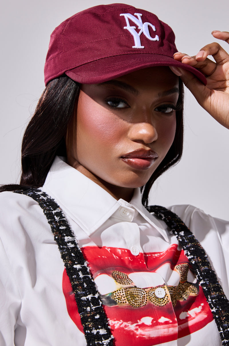 A woman confidently poses in a CITY GURL HAT, white shirt with a bold red lips graphic, and black suspenders, holding the brim of her hat.