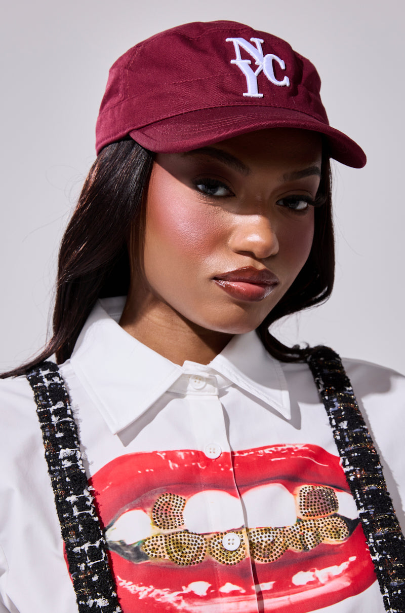 A woman in a burgundy CITY GURL HAT, white graphic lip and gold grills shirt, and black embellished suspenders poses confidently against a plain background.
