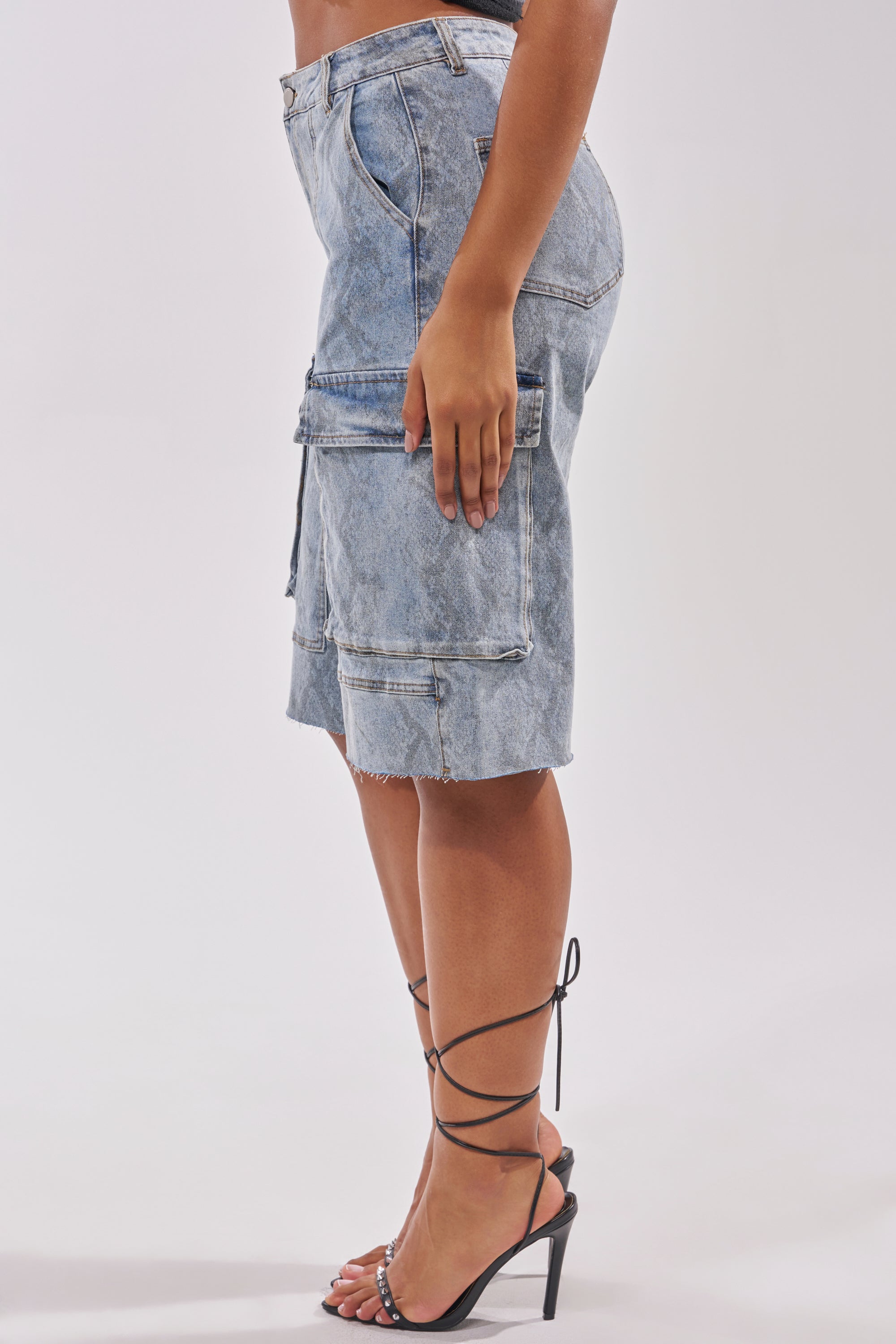 A person models the LITTLE SNAKE BERMUDA SHORT, a light blue denim cargo skirt with a high-rise fit, frayed hem, and large side pockets, paired with black strappy high-heeled sandals. Side photo; plain background. 1
