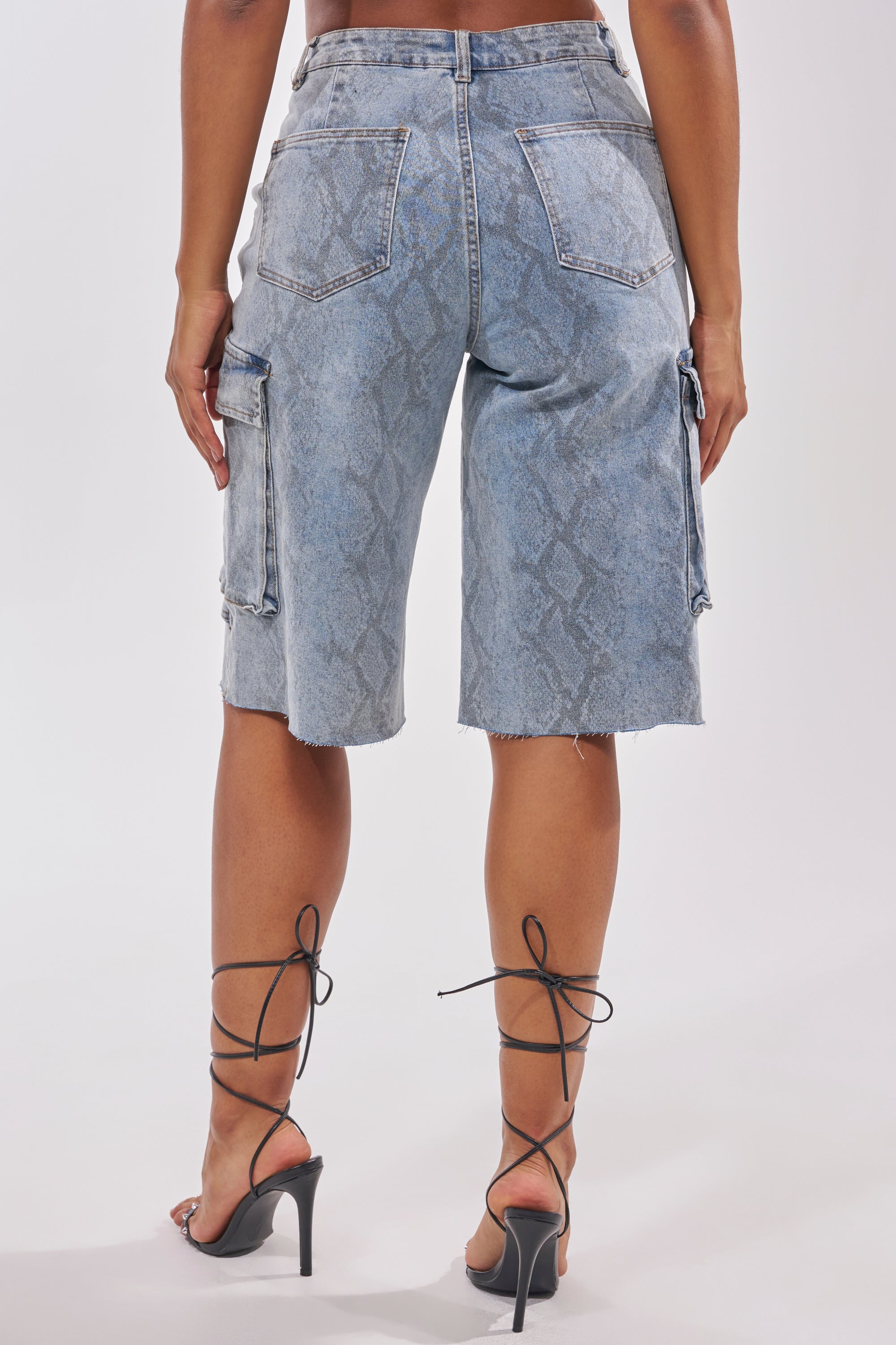A person is shown from the back wearing the LITTLE SNAKE BERMUDA SHORT—high-rise, snake print denim bermuda shorts with frayed hems and side cargo pockets—paired with black lace-up high heel sandals against a plain background. 2
