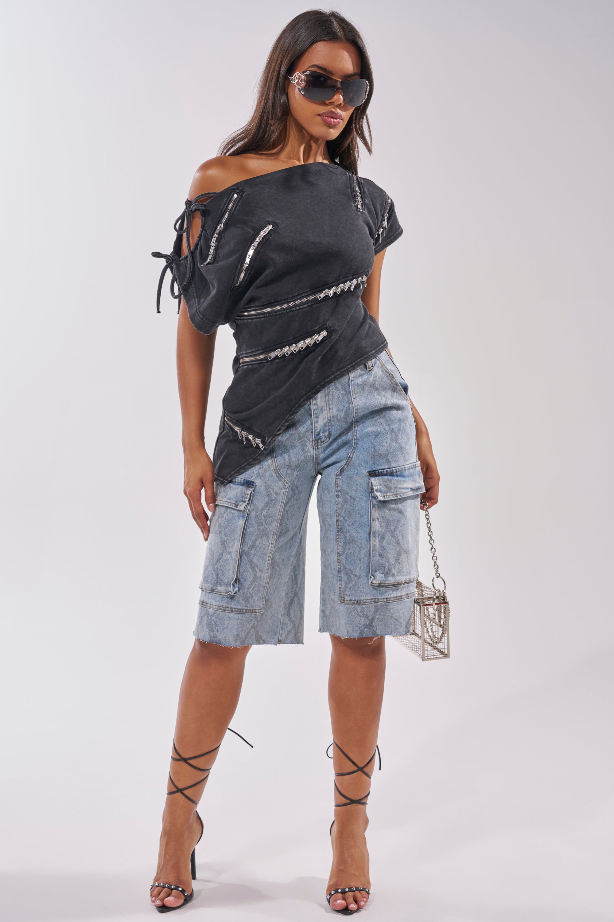 A woman wears the LITTLE SNAKE BERMUDA SHORT in light blue snake print, styled with large sunglasses, a black off-shoulder zippered top, strappy lace-up heels, and a small bag. 3
