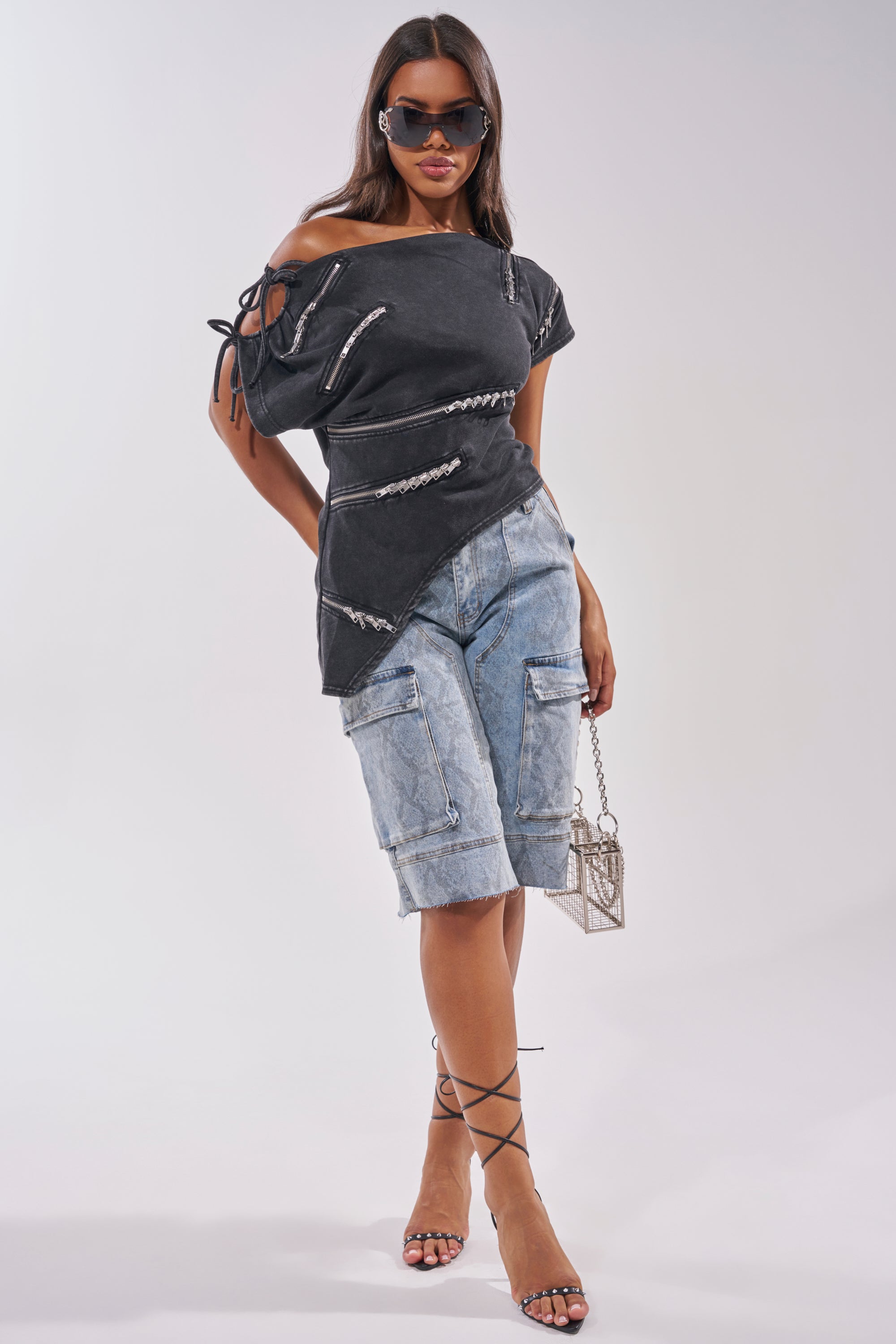 A woman in sunglasses poses stylishly, wearing an asymmetrical black top with silver zippers, LITTLE SNAKE BERMUDA SHORT denim shorts with cargo pockets, strappy high heels, and holding a small metallic handbag. 4