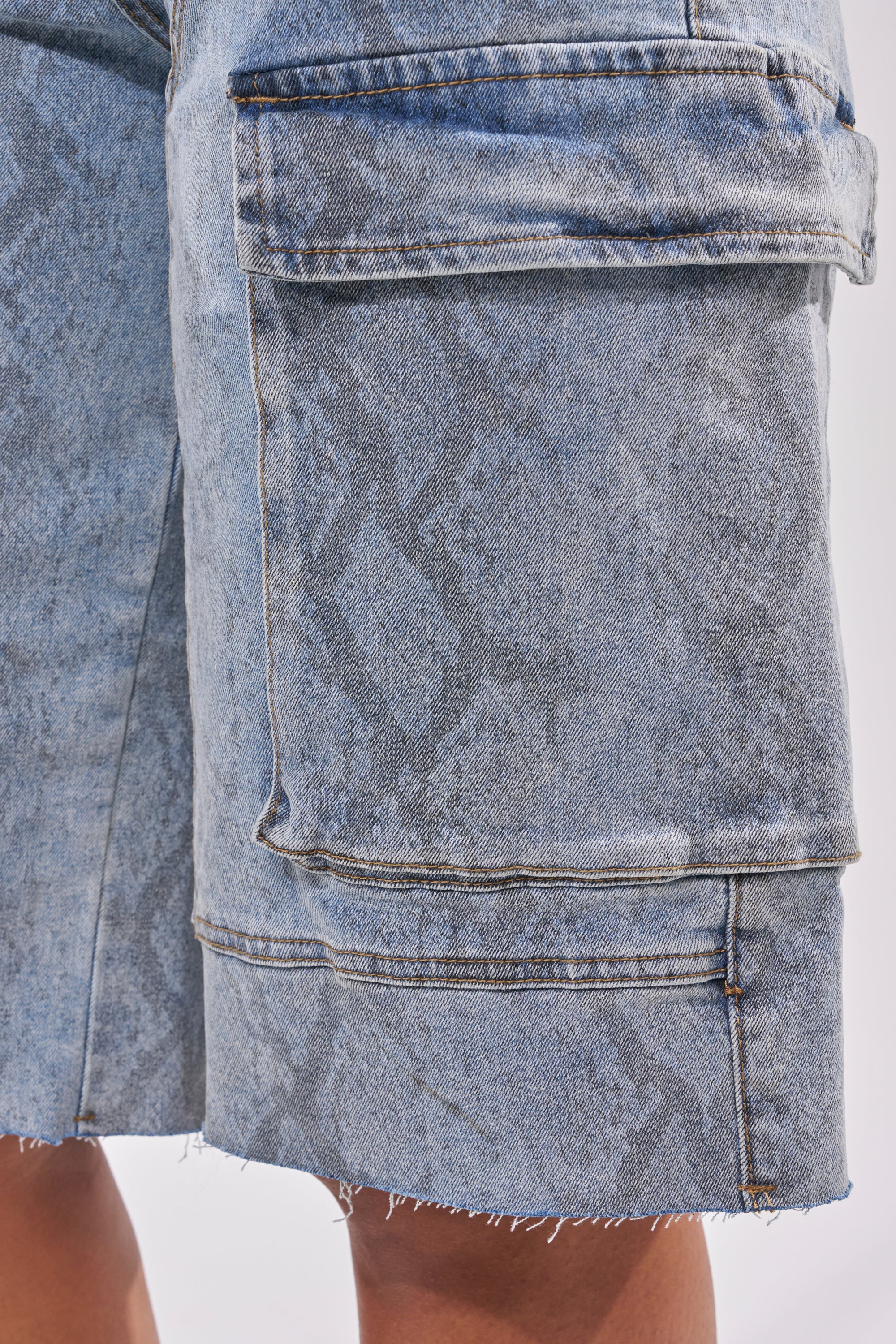 Close-up of a person's leg wearing the LITTLE SNAKE BERMUDA SHORT in light blue denim, featuring a large side pocket, subtle faded patterns, and frayed hems. 6