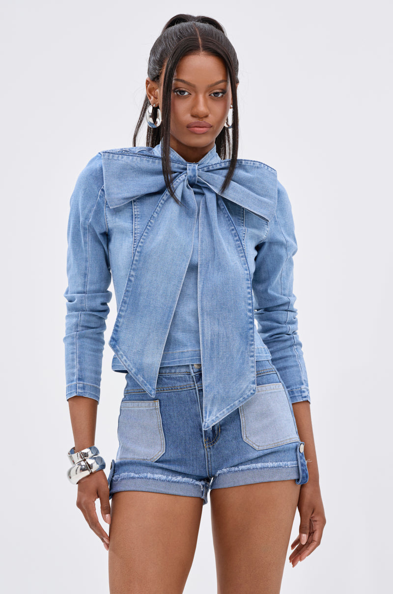 A woman wears the BOW DOWN BISH DENIM LONG SLEEVE TOP, a light blue cropped denim blouse with an oversized bow collar, styled with matching shorts, hoop earrings, silver bracelets, and a high half-ponytail.
