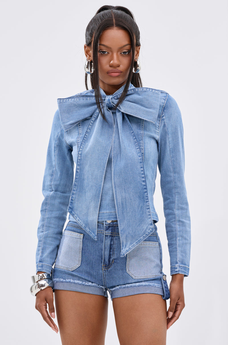 A woman wears the BOW DOWN BISH DENIM LONG SLEEVE TOP, featuring a light blue wash and oversized bow collar, paired with matching cropped high-waisted shorts against a plain light background.