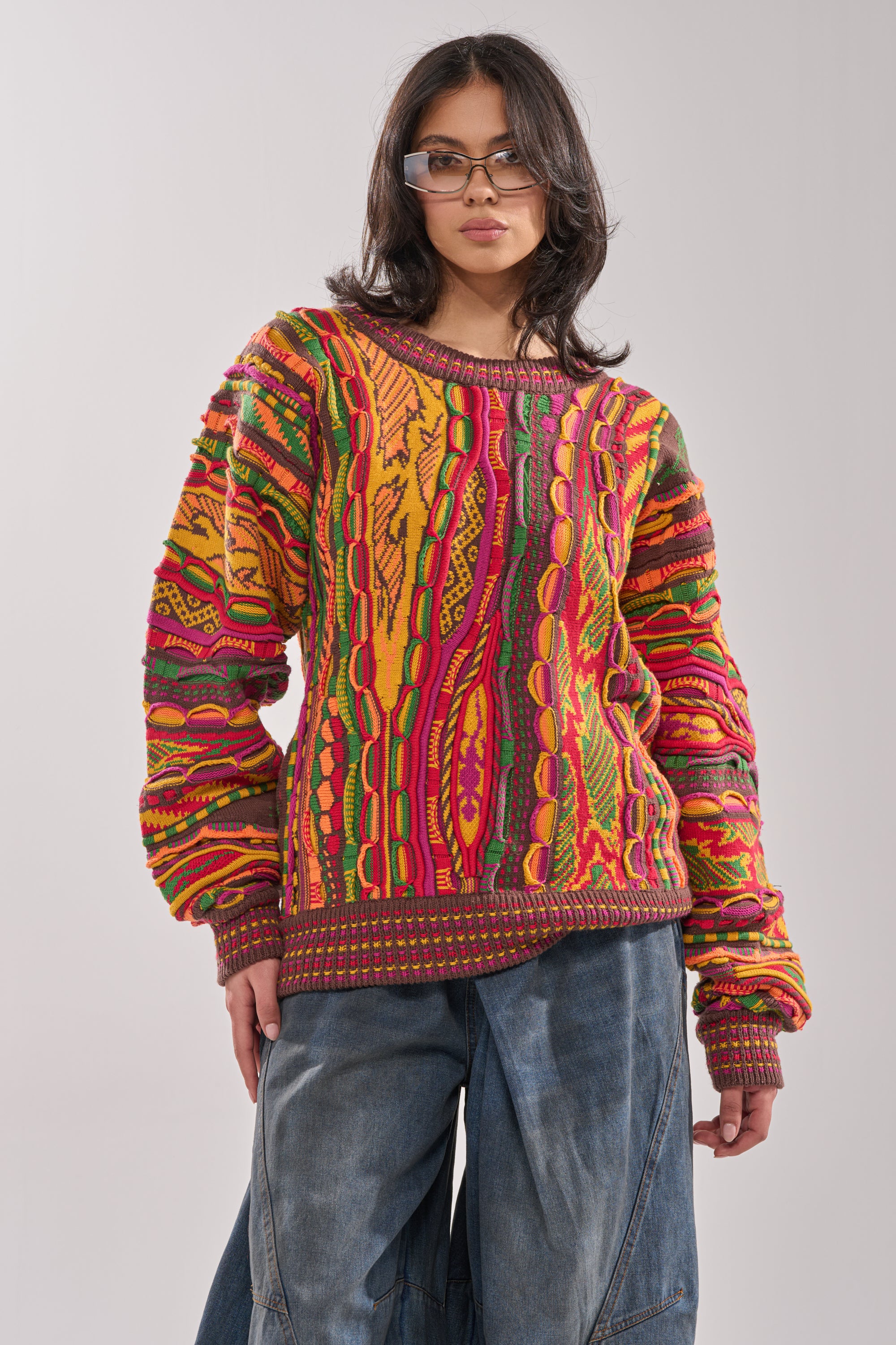 A person wearing glasses, the THROWBACK SPECIAL KNIT SWEATER with bold red, yellow, green, and orange patterns, and loose blue denim jeans stands against a plain light background. 0
