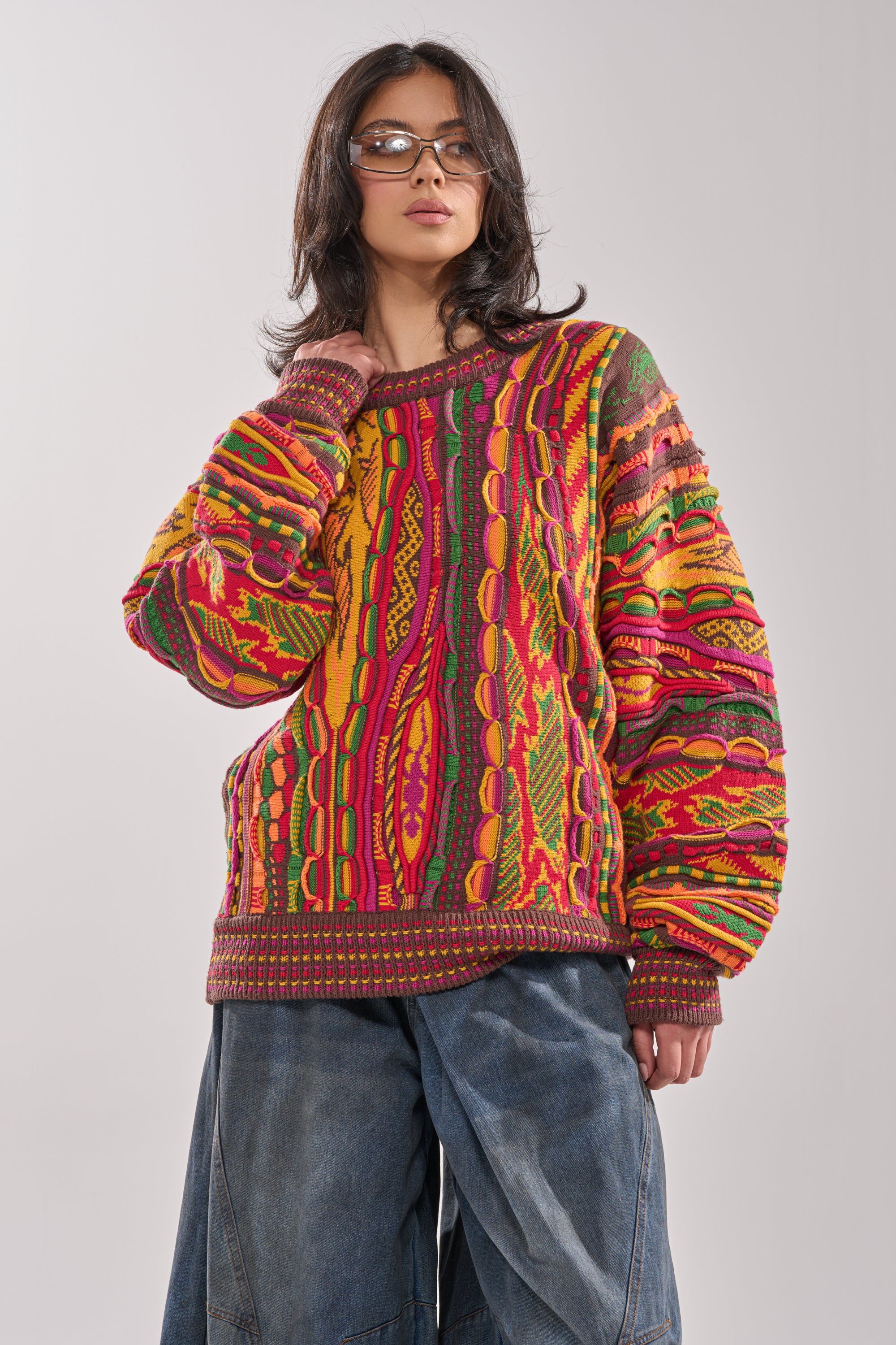 A person wearing glasses, the THROWBACK SPECIAL KNIT SWEATER featuring retro multicolor designs, and wide-leg blue jeans stands against a plain light background. 1