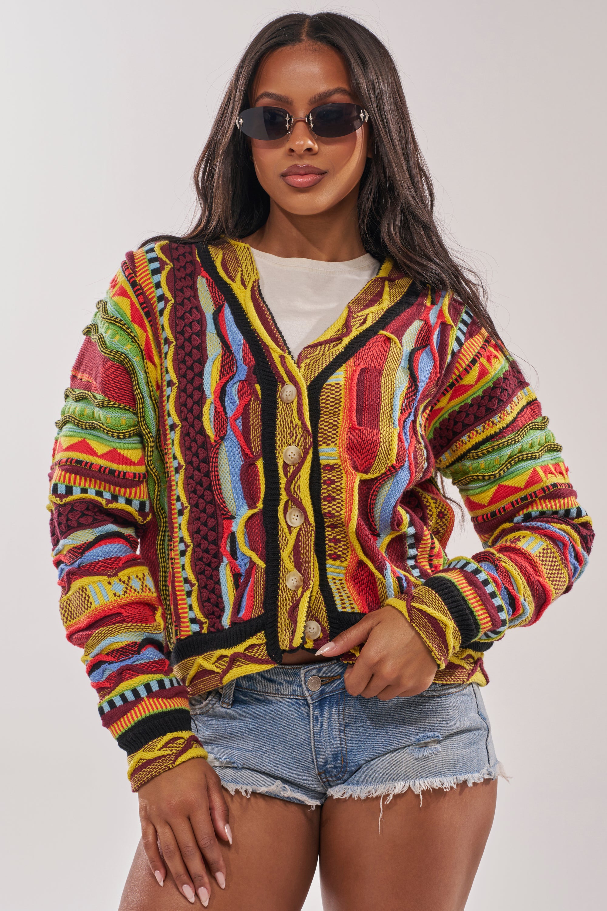 A woman stands confidently against a plain background wearing dark sunglasses, a white top, distressed denim shorts, and the MADE YA LOOK KNIT SWEATER—a colorful vintage-inspired cardigan. 0