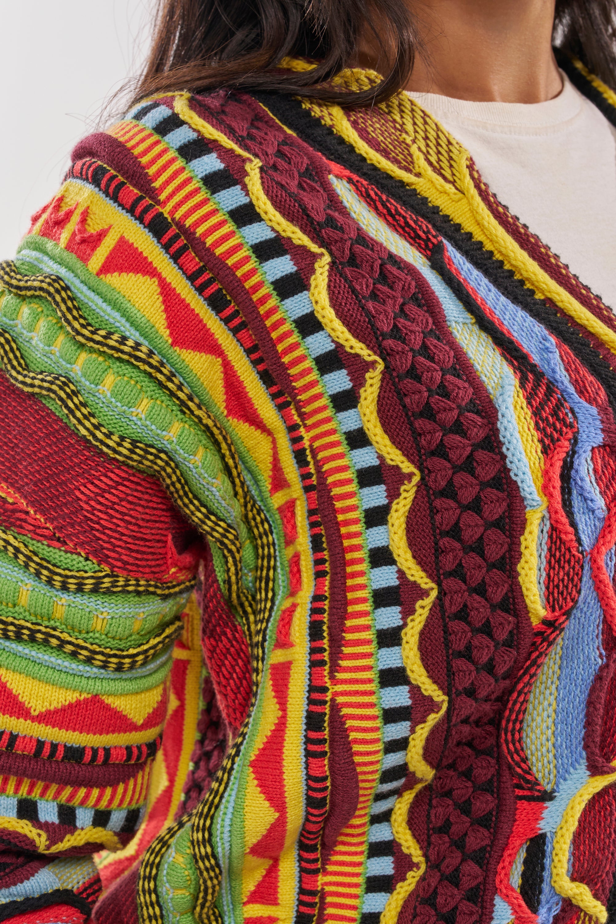 A close-up of someone wearing the MADE YA LOOK KNIT SWEATER, a colorful knit featuring bold red, yellow, green, blue, and black patterns and textured designs as the person faces slightly away from the camera. 9