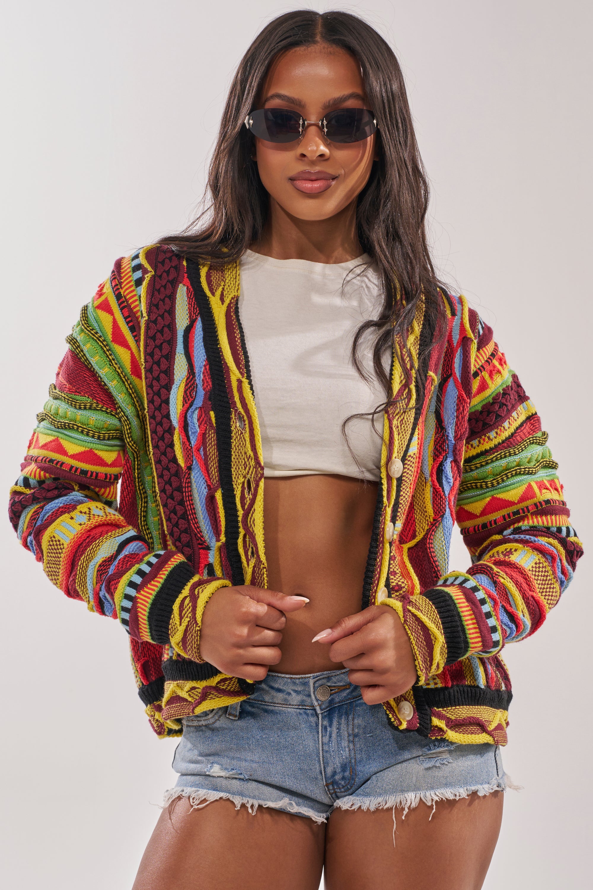 Woman wearing dark sunglasses, a cropped white top, denim shorts, and the MADE YA LOOK KNIT SWEATER with multicolored patterns, posing confidently against a plain background. 1