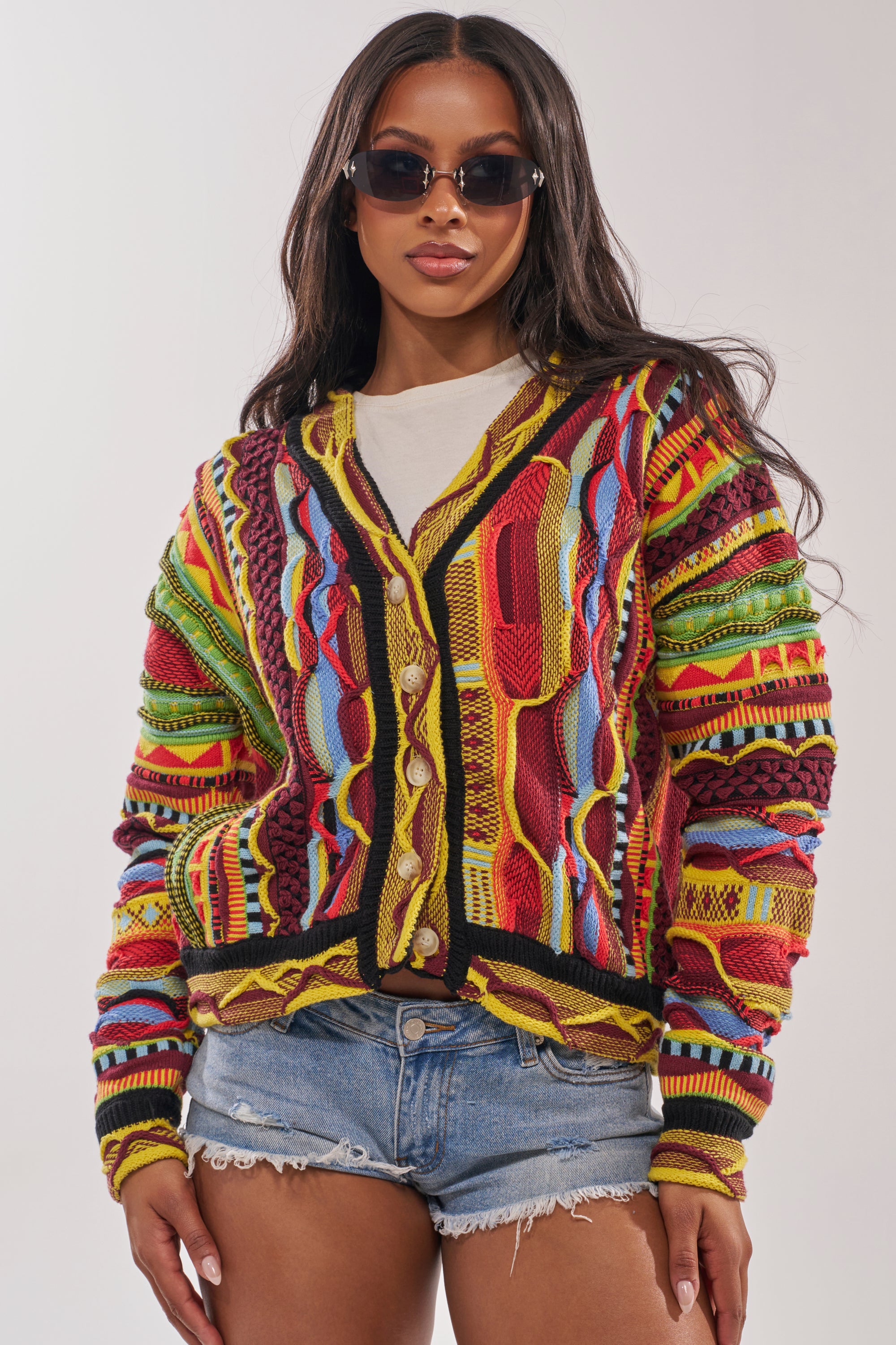 A woman wearing sunglasses, the MADE YA LOOK KNIT SWEATER with vibrant patterns, a white shirt, and distressed denim shorts stands against a plain background. 2