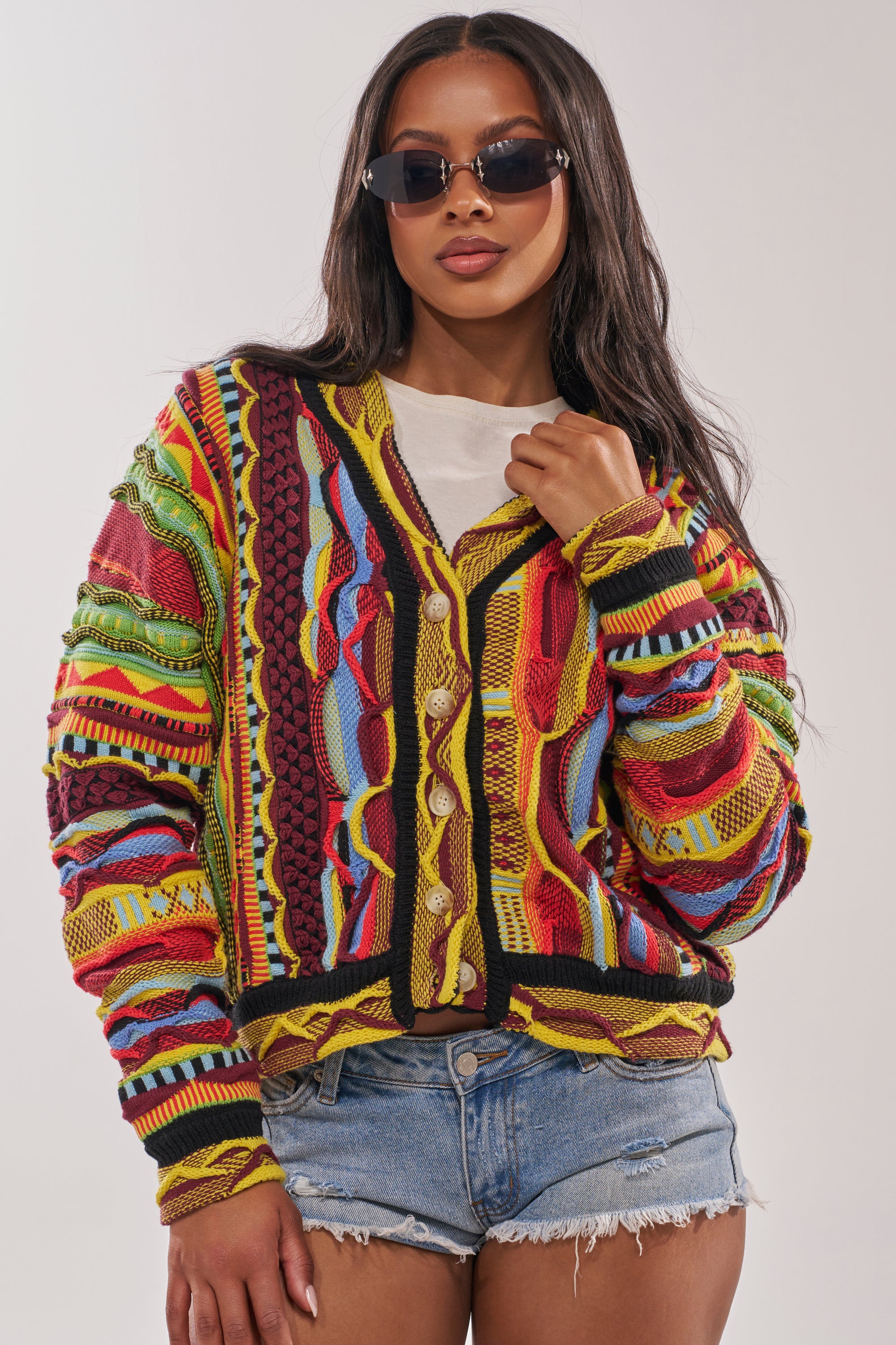 A woman poses confidently against a plain background wearing the MADE YA LOOK KNIT SWEATER over a white shirt, denim shorts, and dark sunglasses. 3