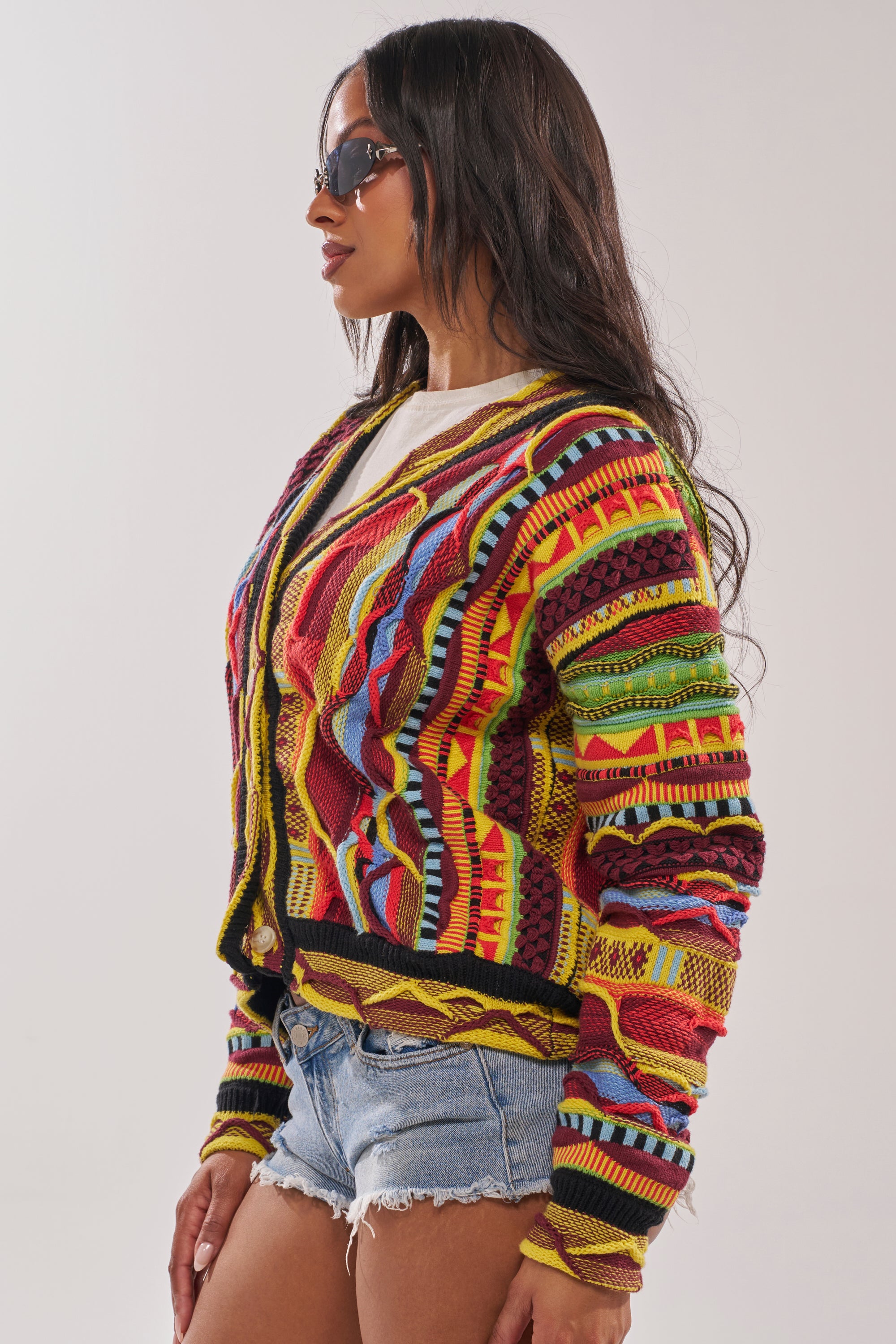 Woman wears the MADE YA LOOK KNIT SWEATER over a white shirt with denim shorts and sunglasses, standing sideways against a plain light background. 4