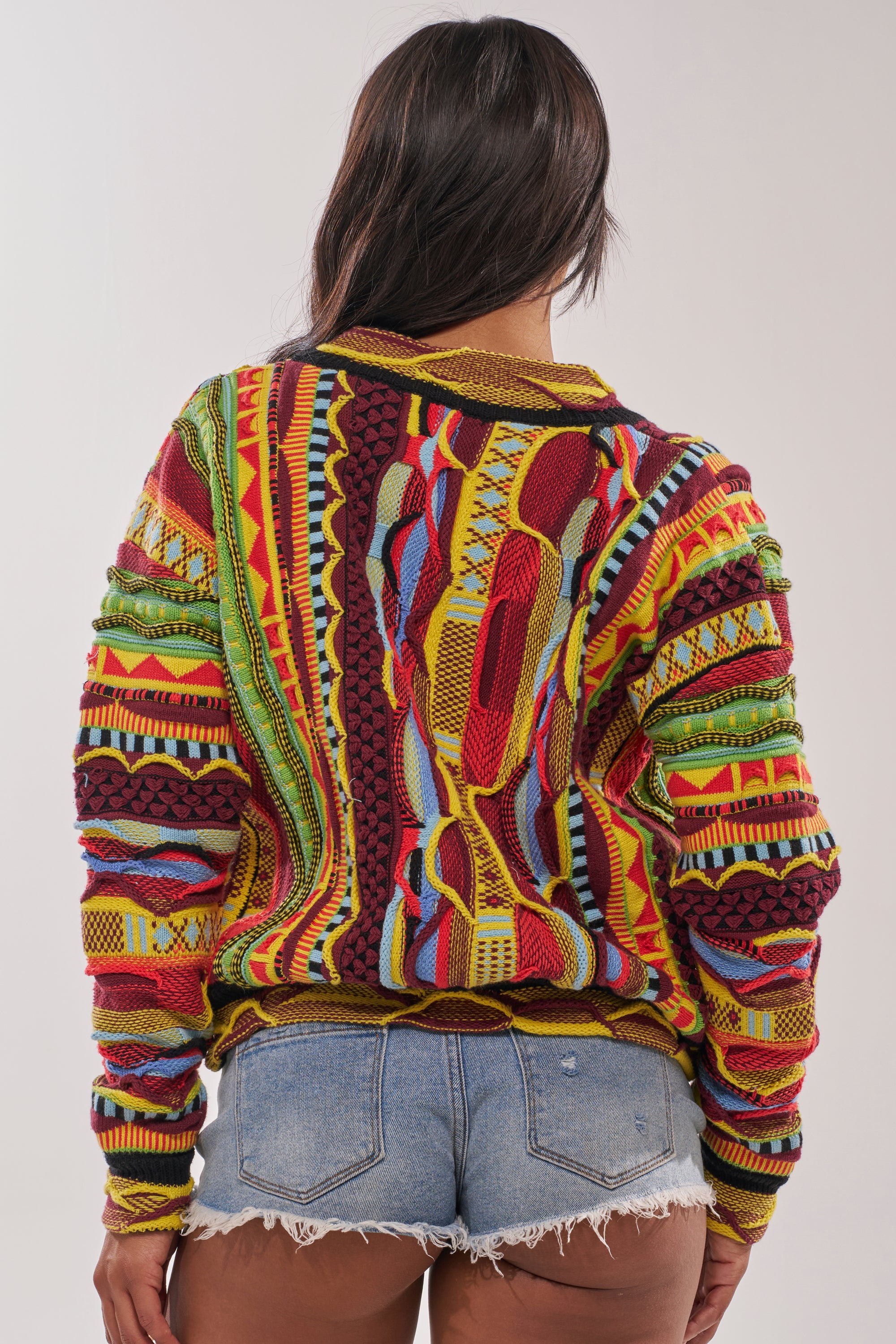 A person with long dark hair, seen from behind, wears the MADE YA LOOK KNIT SWEATER featuring colorful vintage designs in red, yellow, green, and blue, paired with light blue denim shorts. 5