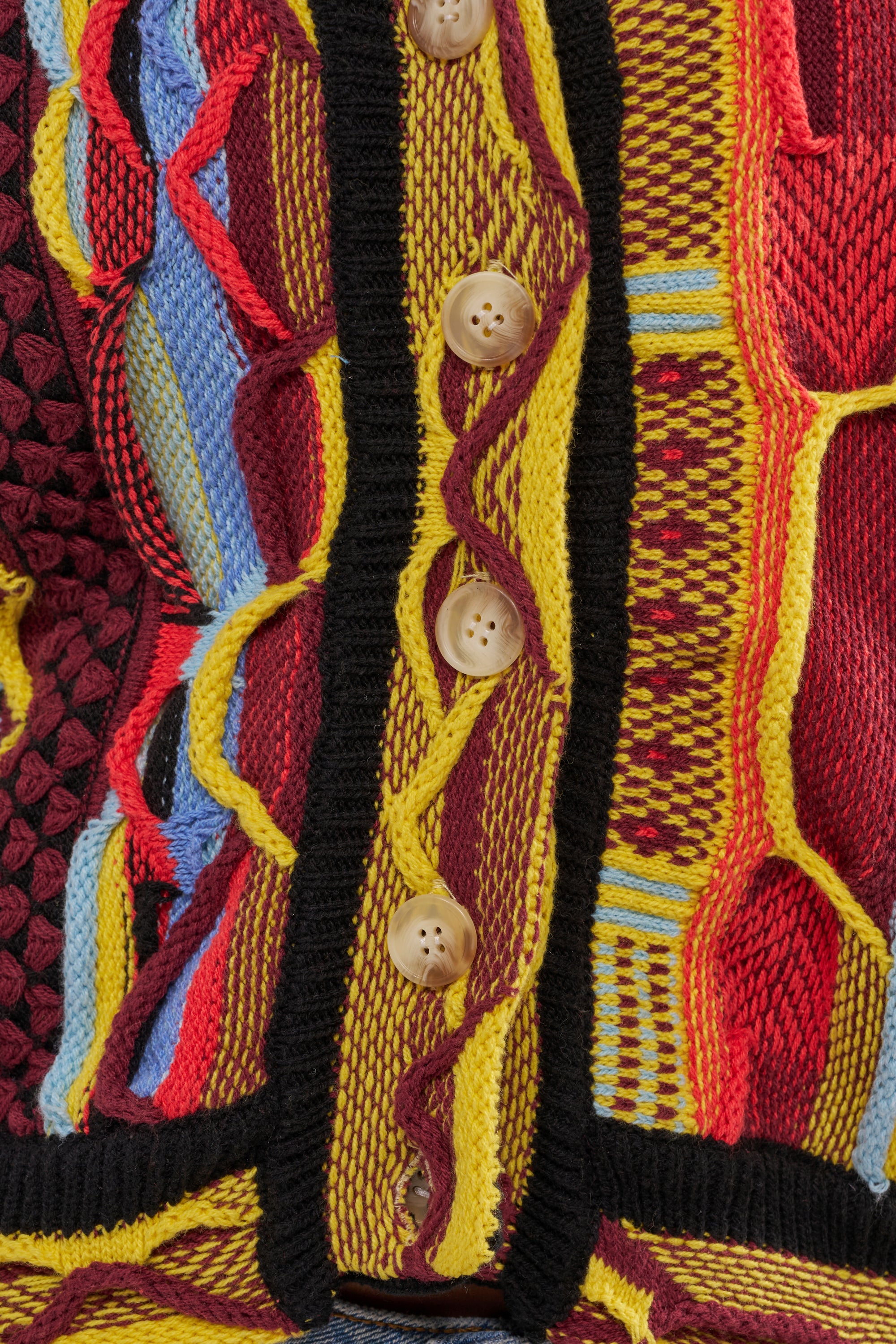 Close-up of the MADE YA LOOK KNIT SWEATER, featuring wavy patterns in yellow, red, blue, and brown with black trim along the center and a row of tan buttons. 8