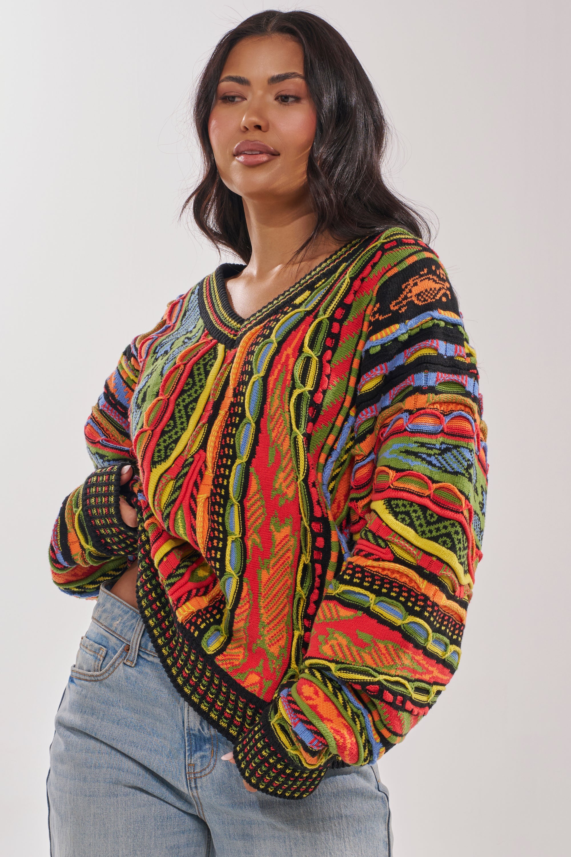 A woman with long dark hair wears the COSMIC WEAVE V-NECK SWEATER, featuring bold multicolor patterns and loose sleeves, paired with light blue jeans against a plain light background.