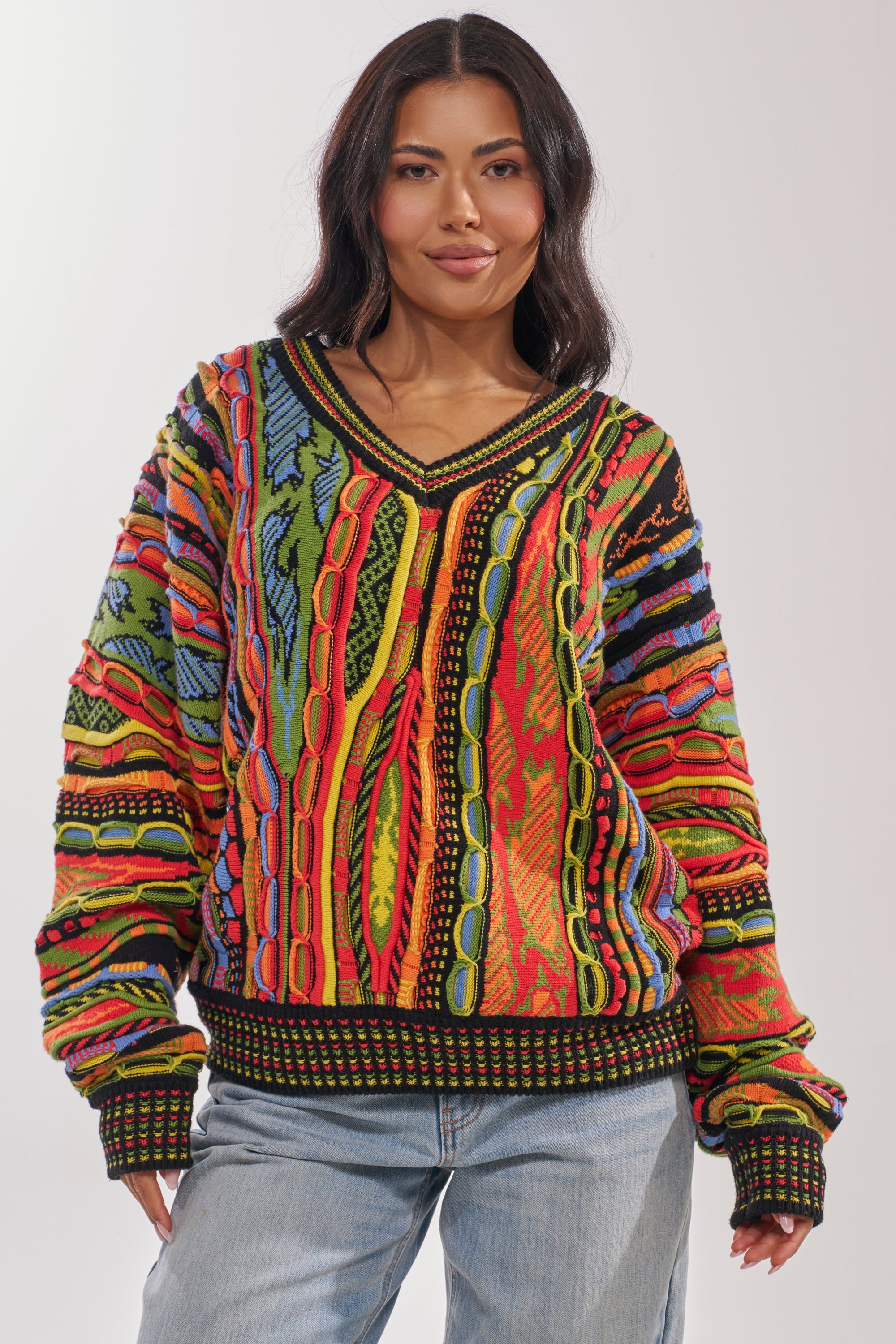 A woman smiles against a plain background in the COSMIC WEAVE V-NECK SWEATER, an oversized piece featuring bold, abstract red, yellow, blue, and green designs, paired with light blue jeans. 1